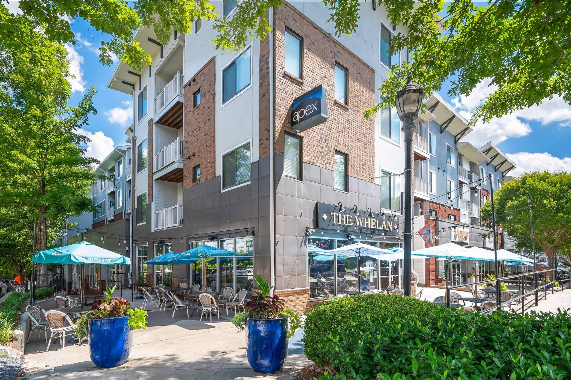 Modern apartment building with ground-floor restaurant, outdoor seating, blue umbrellas, and leafy trees lining the street.