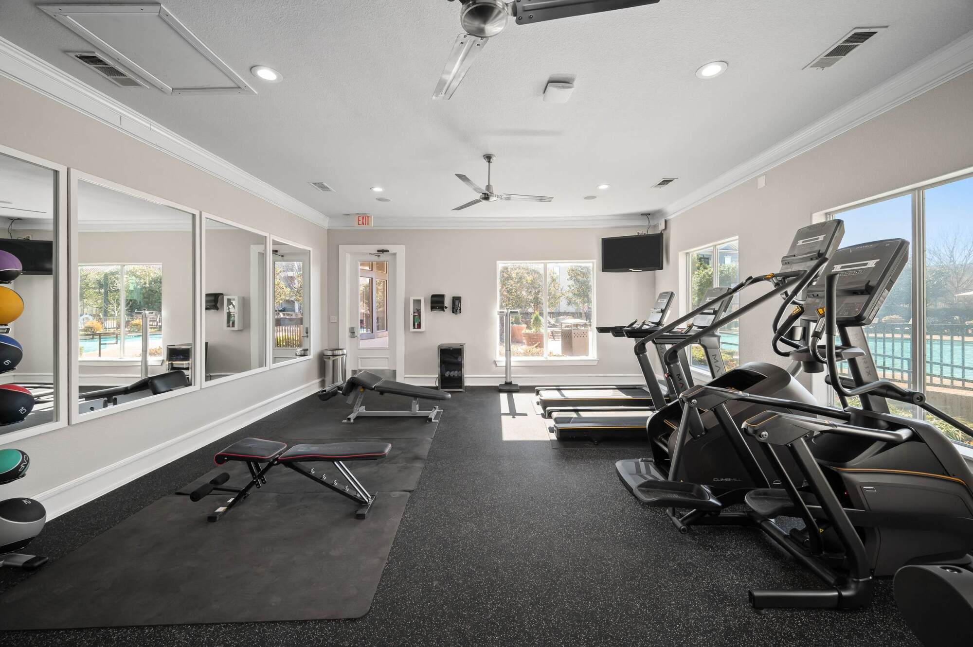 Modern gym room with treadmills, elliptical machines, weights, mirrors, and pool view through large windows.
