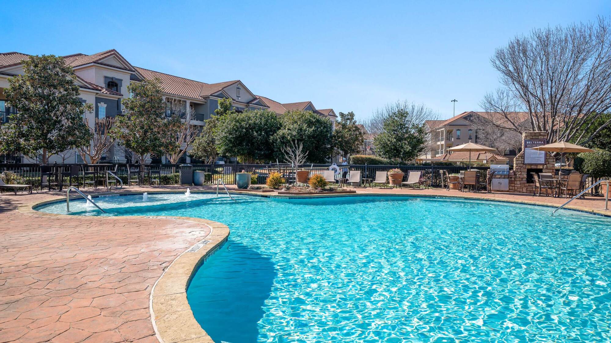 Outdoor swimming pool with lounge chairs and umbrellas, surrounded by apartment buildings and trees, on a sunny day.