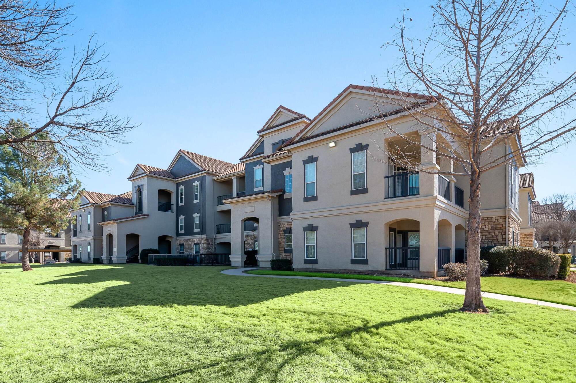 Modern apartment buildings with balconies and large windows, facing a sunny, grassy lawn with a few trees.