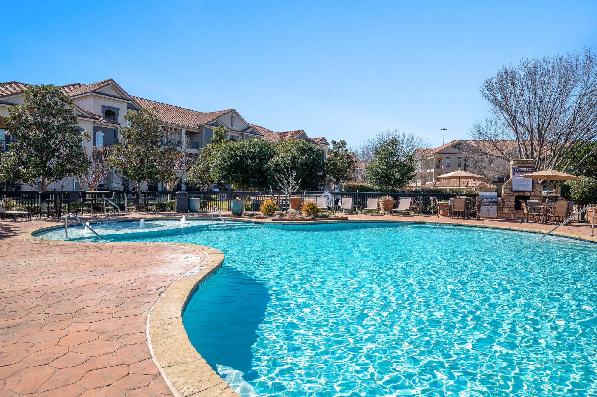 Outdoor swimming pool surrounded by lounge chairs and trees near apartment buildings on a sunny day.