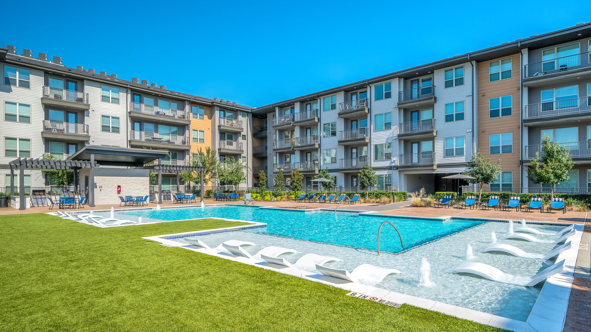 Modern apartment complex with a large outdoor pool, lounge chairs, and landscaped green area under a clear sky.