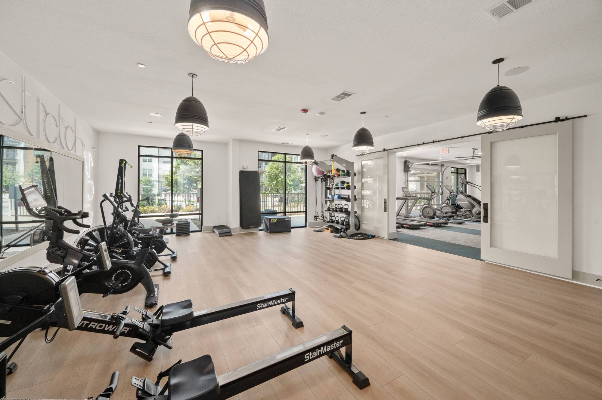Modern gym with rowers, bikes, weights, large windows, and light wood flooring, filled with natural light.