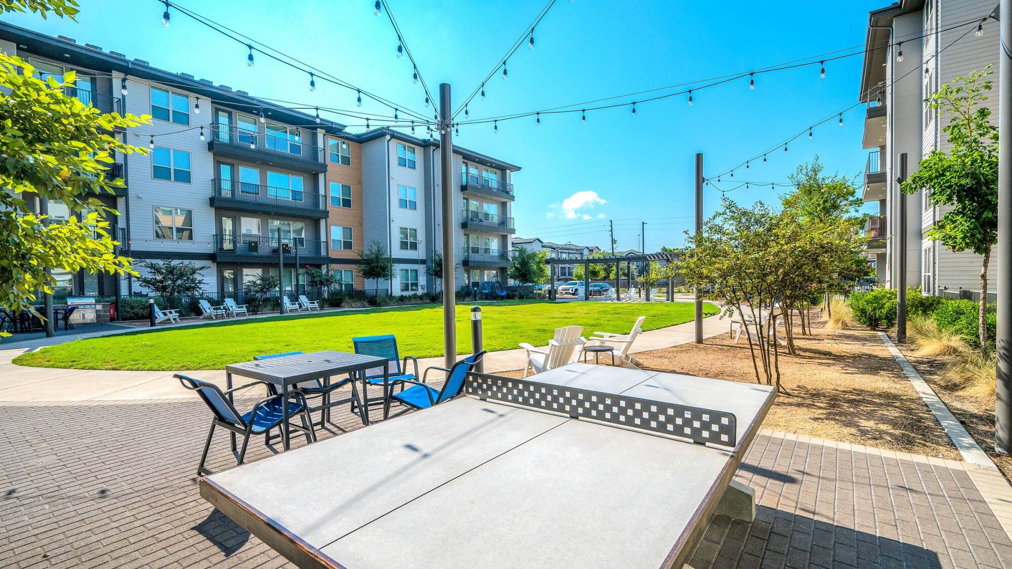 Outdoor courtyard with a ping pong table, seating areas, string lights, grass, and modern apartment buildings.