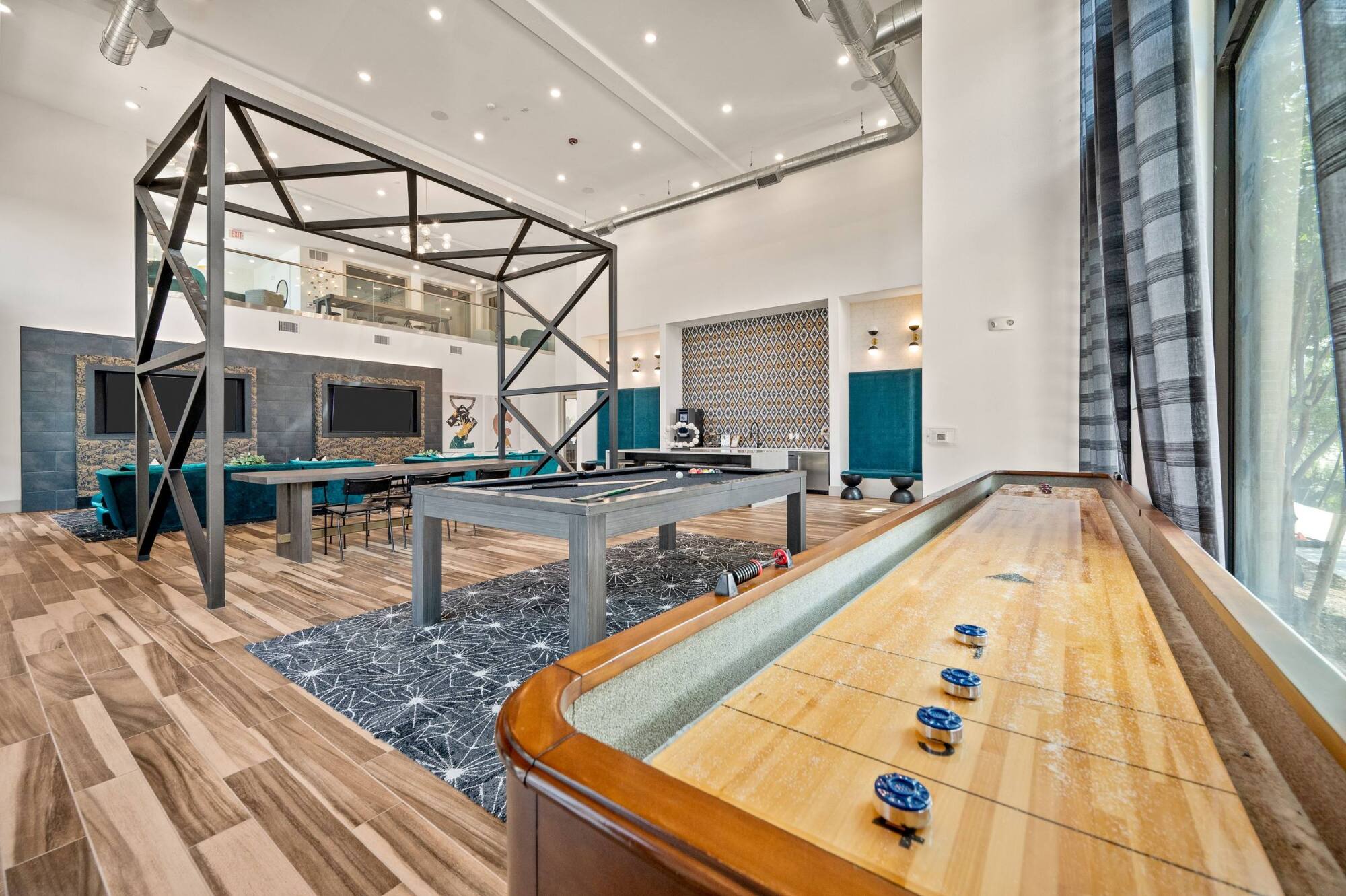 Modern game room with shuffleboard, billiards table, TVs, and stylish decor featuring wood floors and large windows.