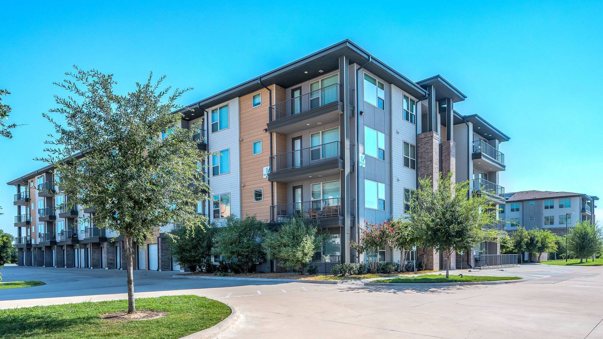 Modern four-story apartment building with balconies, surrounded by trees and a wide driveway under a clear blue sky.