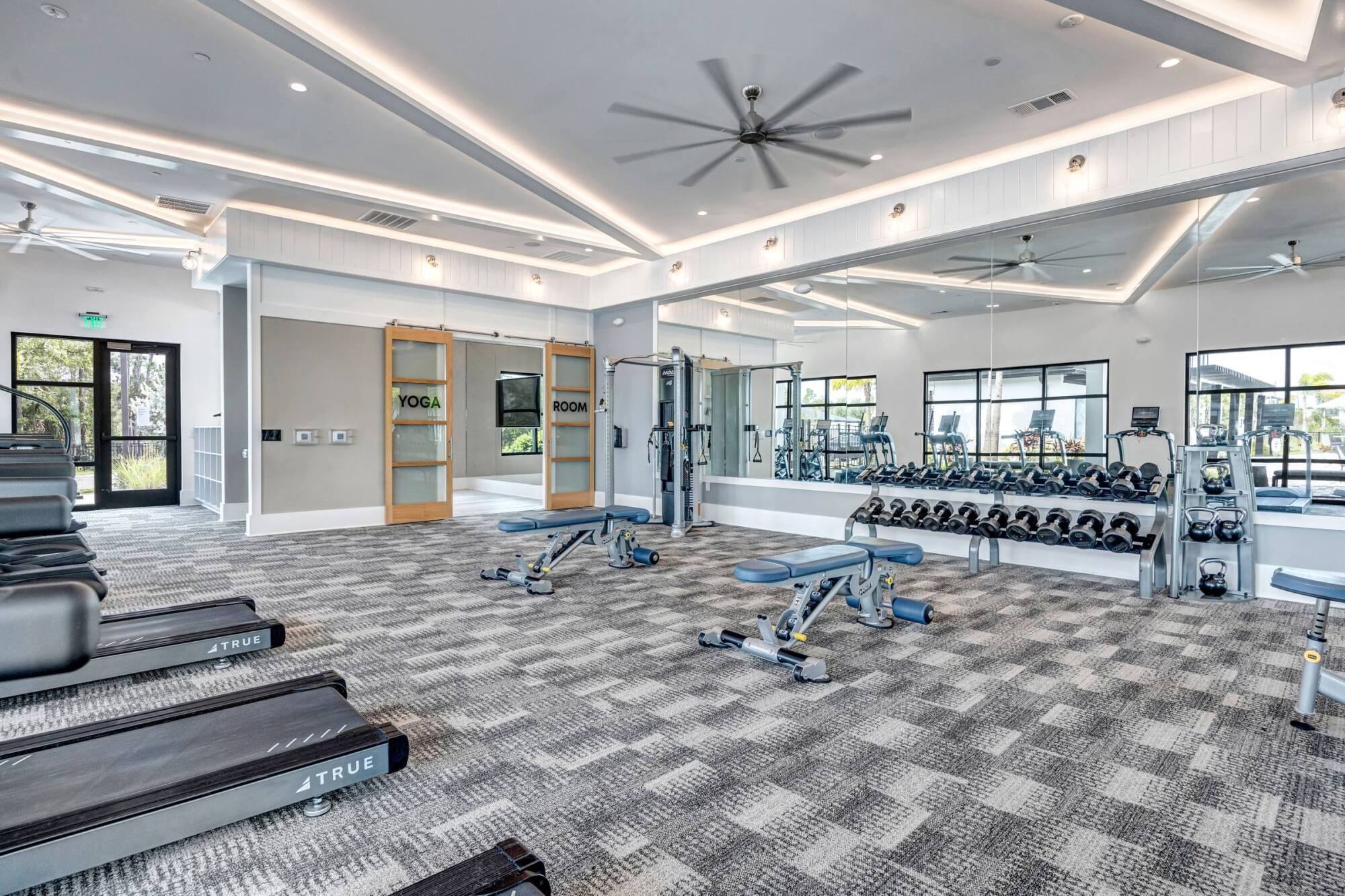Modern gym with treadmills, benches, dumbbells, mirrored wall, and yoga room with glass doors.