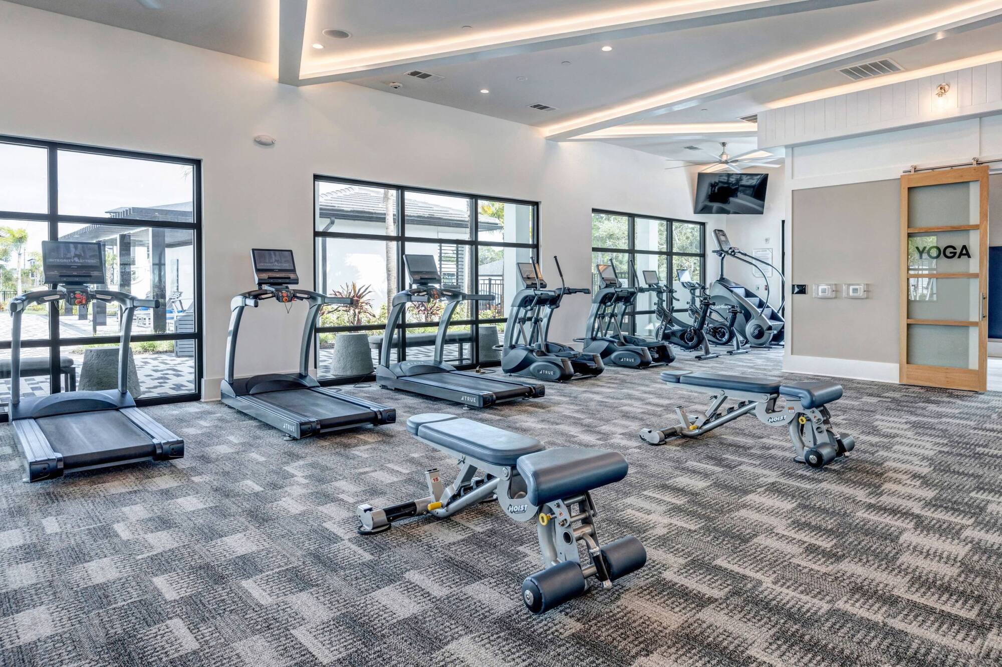 Modern gym with treadmills, ellipticals, benches, large windows, and a yoga room door in the background.