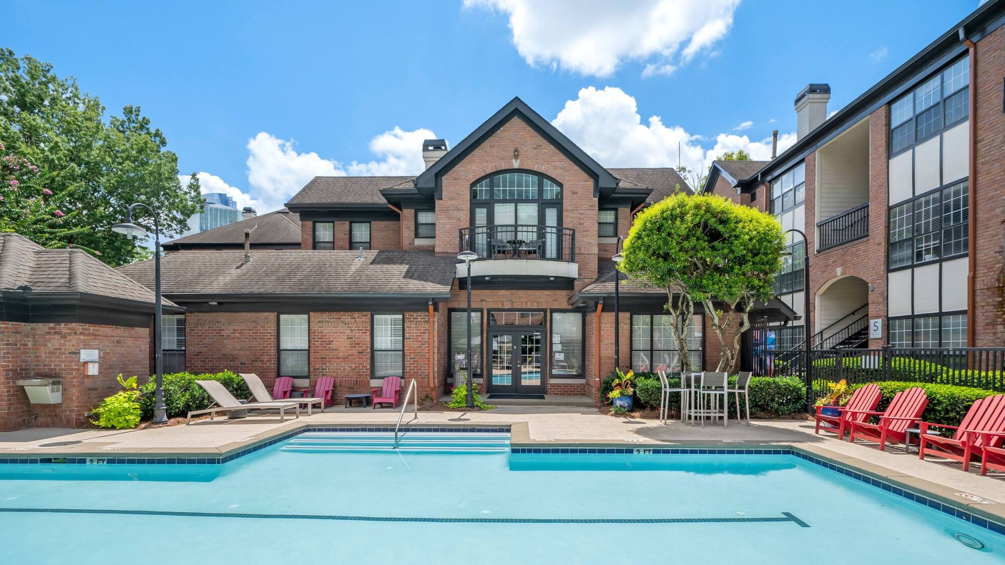 Outdoor swimming pool with lounge chairs and red Adirondack chairs in front of a brick apartment building.