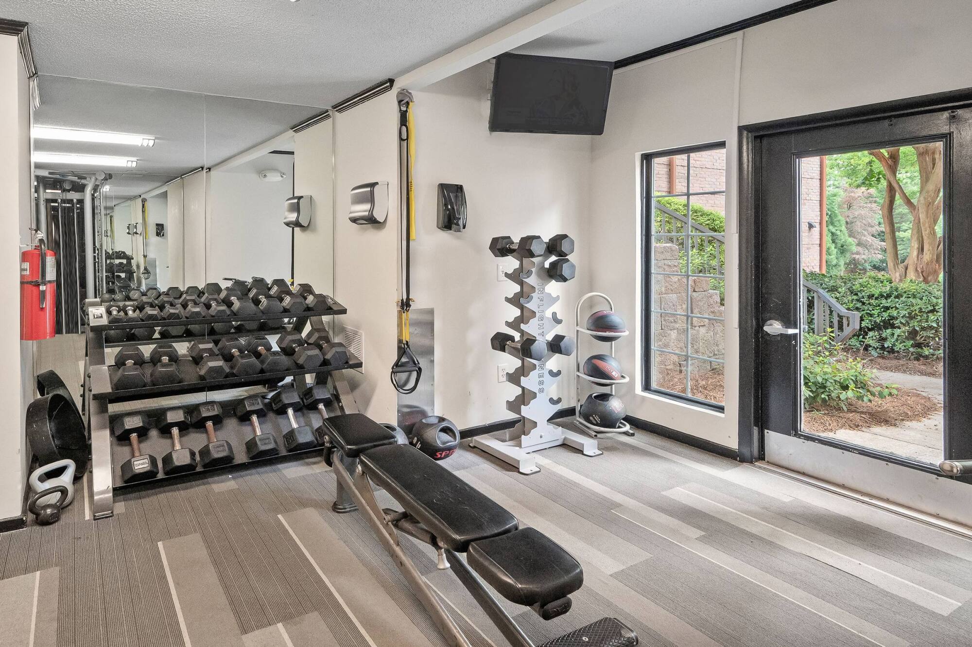Small gym room with free weights, bench, exercise balls, wall mirror, TV, and large window facing greenery.