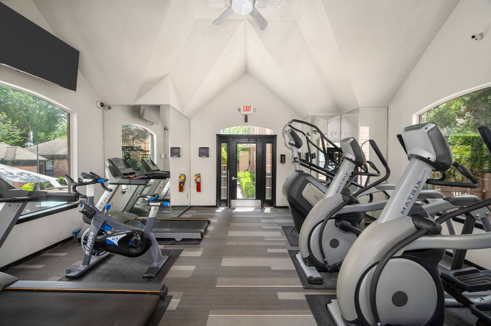 Small gym with treadmills, exercise bikes, and ellipticals, featuring large windows and natural light.