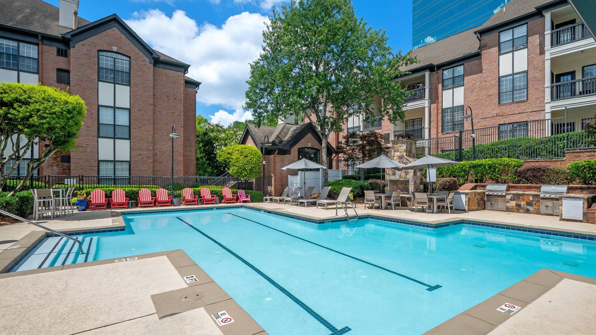 Apartment complex pool area with lounge chairs, umbrellas, and barbecue grills on a sunny day.