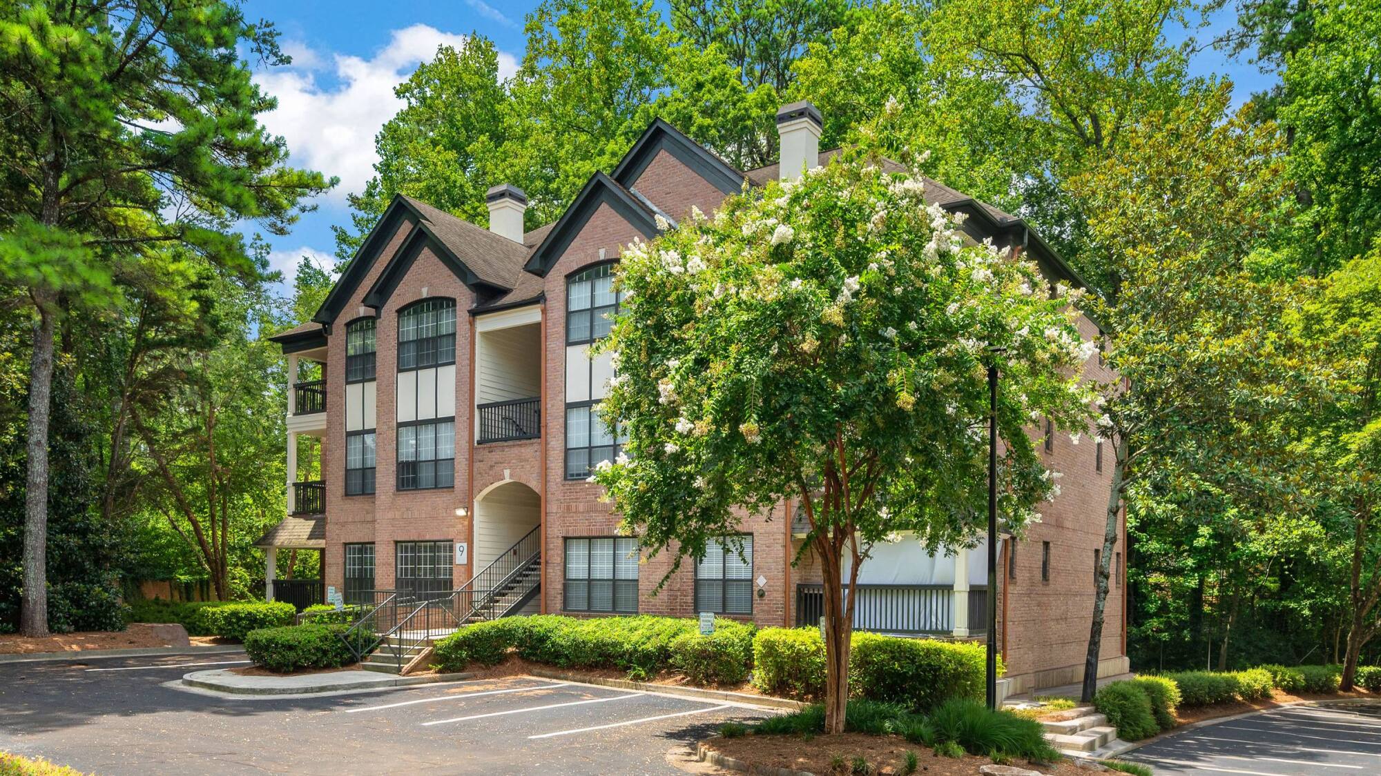 Two-story brick apartment building with large windows, surrounded by trees, next to an empty parking lot.