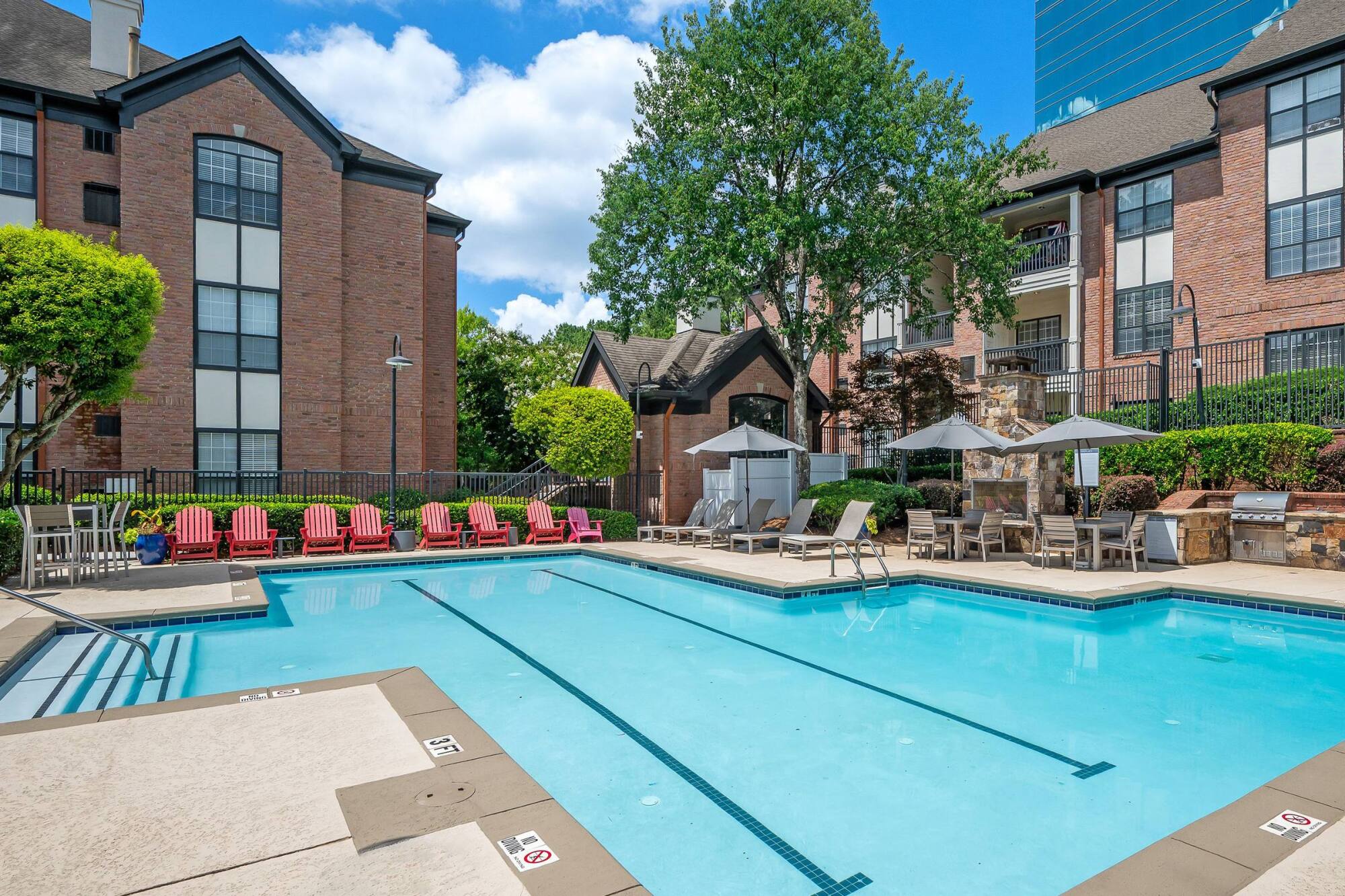 Outdoor apartment pool with red chairs, lounge seating, umbrellas, and surrounding brick buildings on a sunny day.
