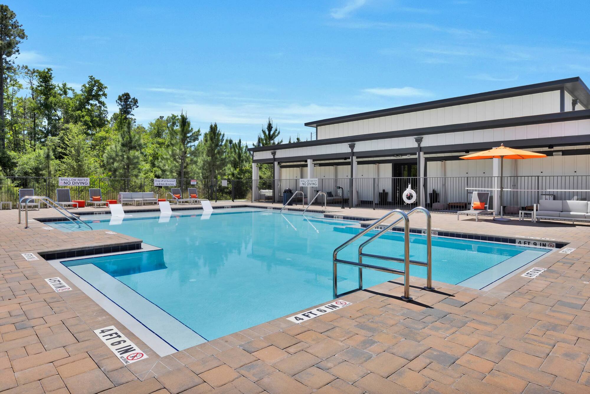 An outdoor residential swimming pool area with lounge chairs, white modern buildings, and clear blue skies.