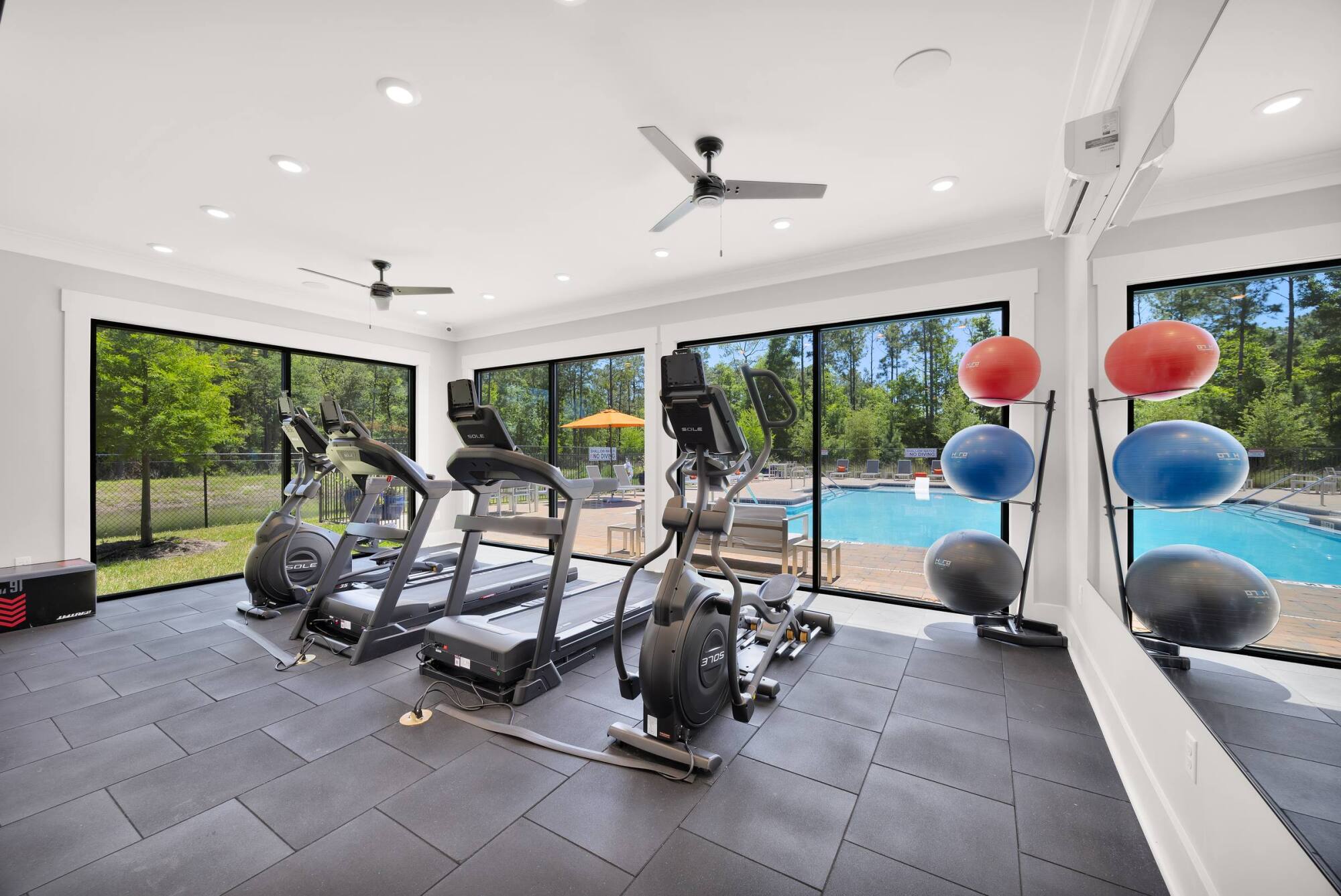 A modern fitness center with treadmills and elliptical machines facing large windows that offer a view of the outdoor swimming pool.
