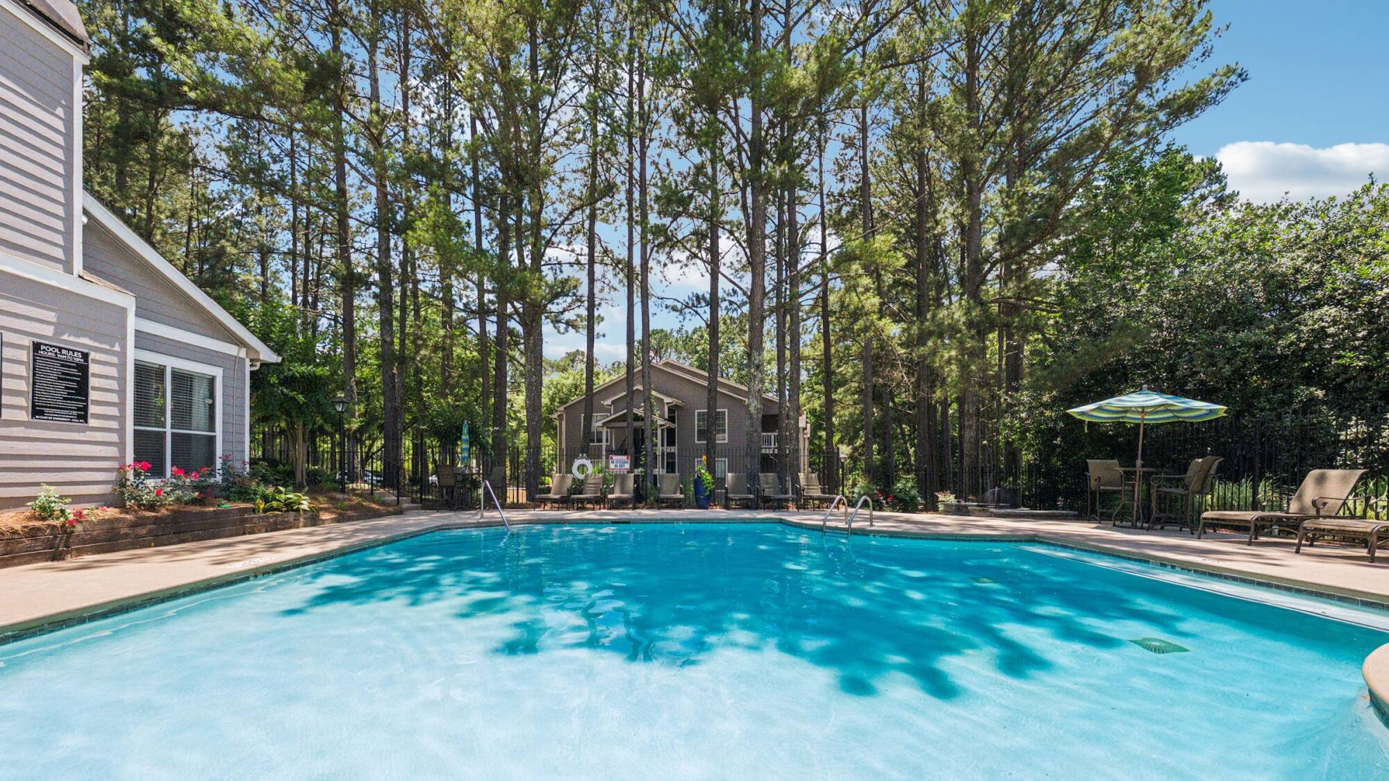 Large backyard pool surrounded by tall trees, patio chairs, and a house in the background on a sunny day.