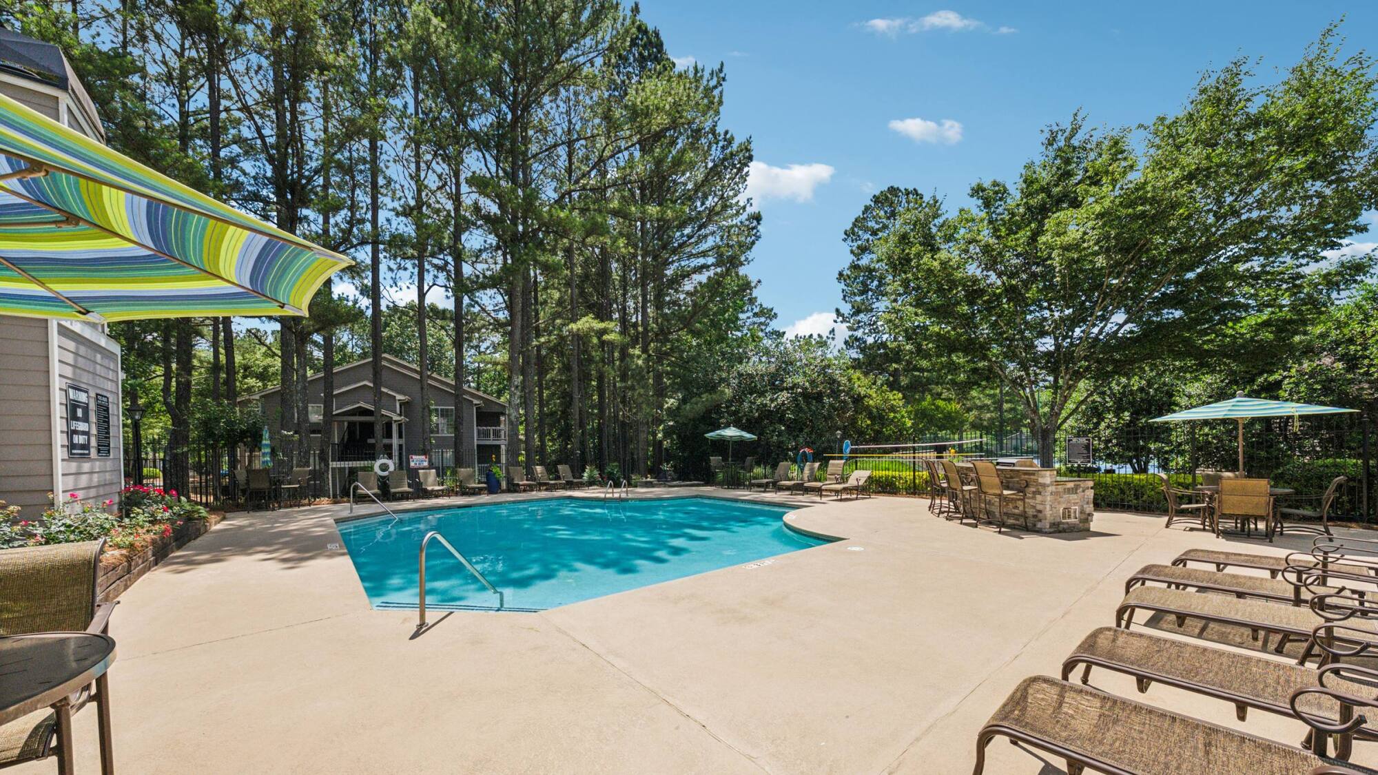 Outdoor swimming pool surrounded by lounge chairs, tables, umbrellas, and tall trees on a sunny day.