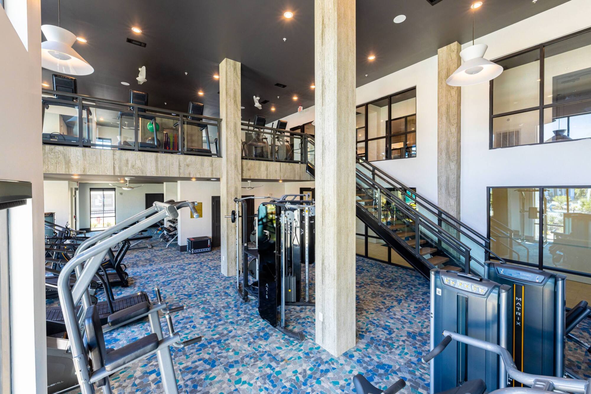 Modern gym with various exercise machines, tall columns, and an upper-level glass balcony overlooking the space.