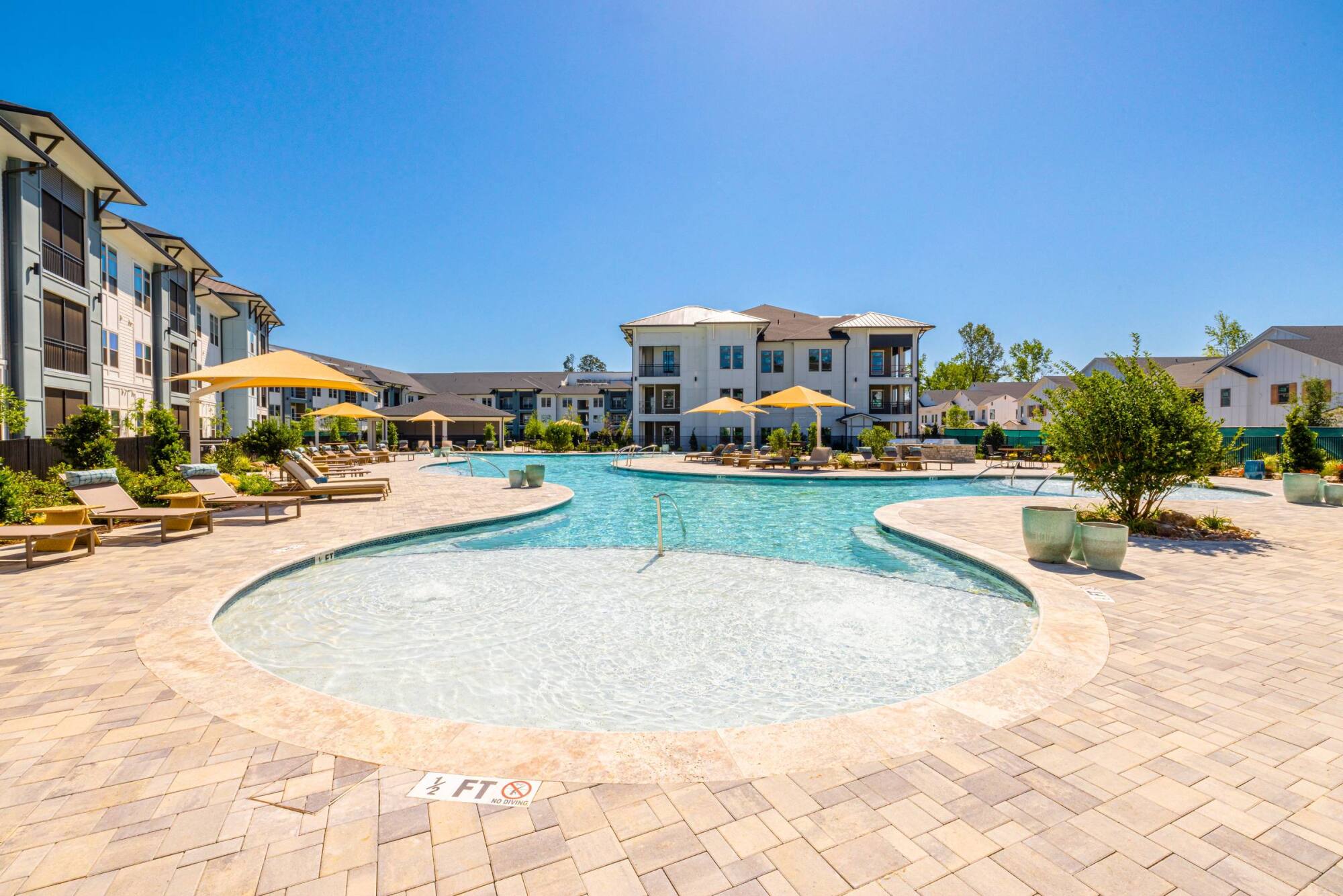 Sunny resort-style pool area with lounge chairs, umbrellas, and surrounding modern apartment buildings.