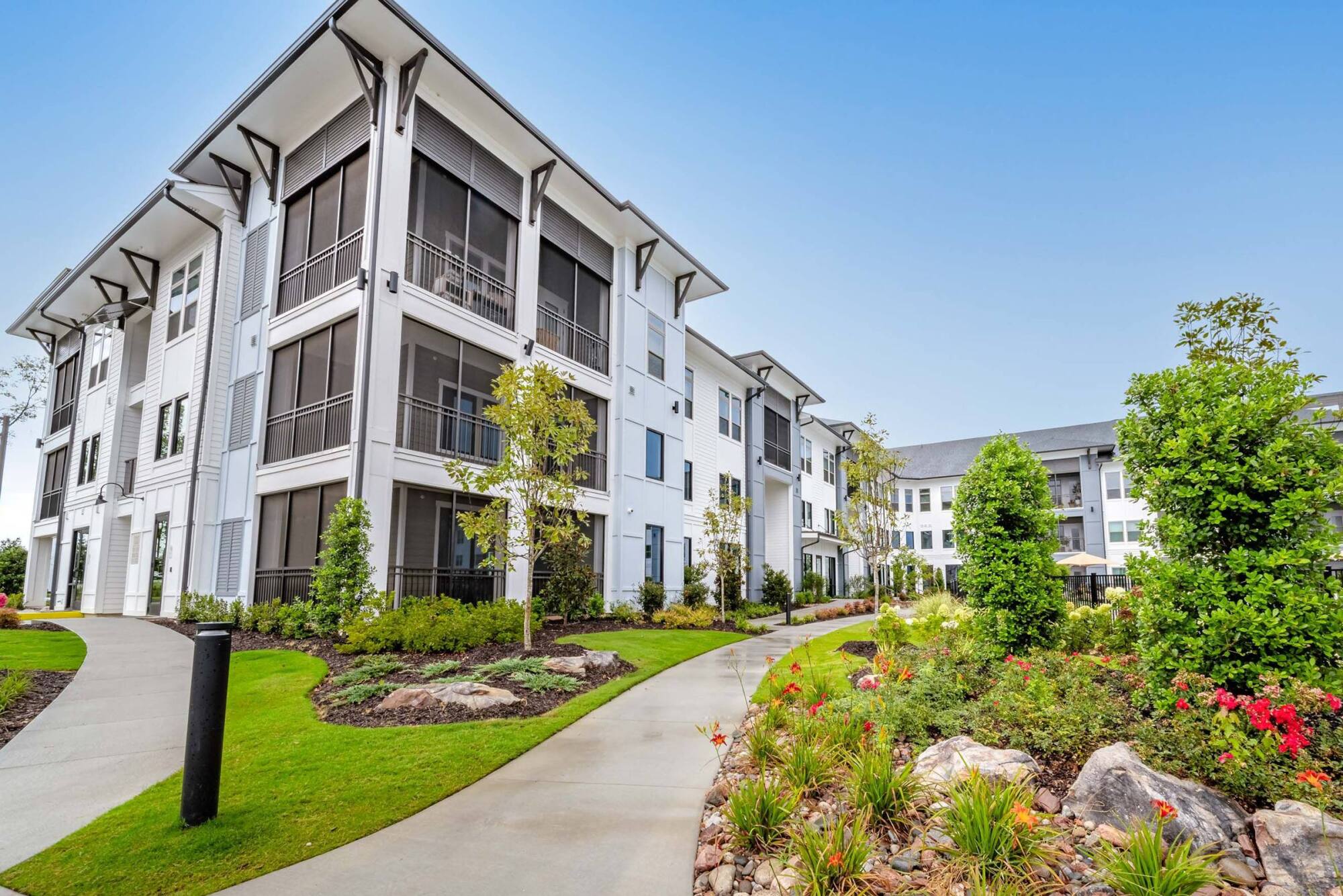 Modern white apartment buildings with large windows, landscaped greenery, and winding walkways under a clear sky.