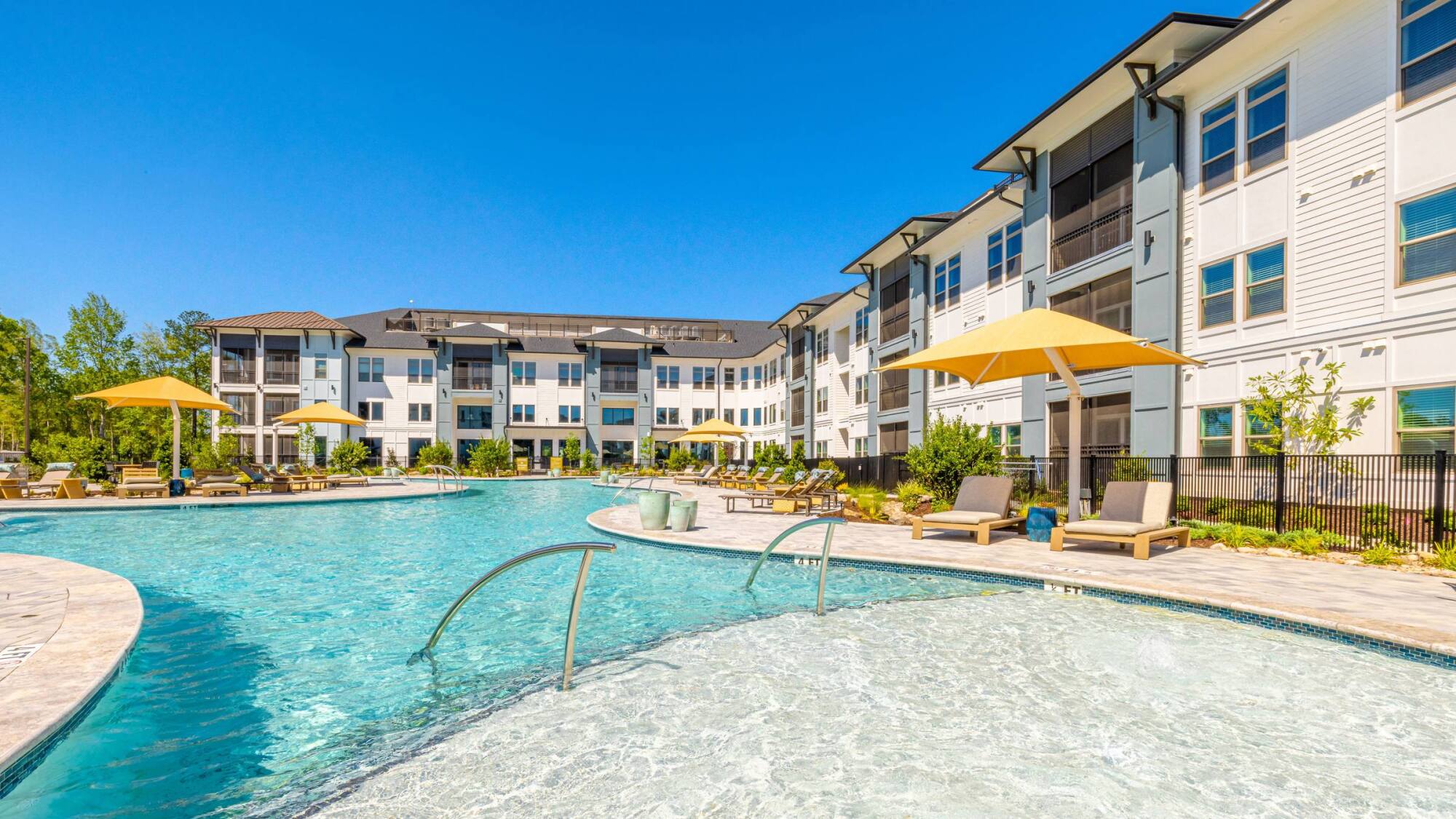 Modern apartment complex with a large outdoor pool, lounge chairs, and yellow umbrellas on a sunny day.
