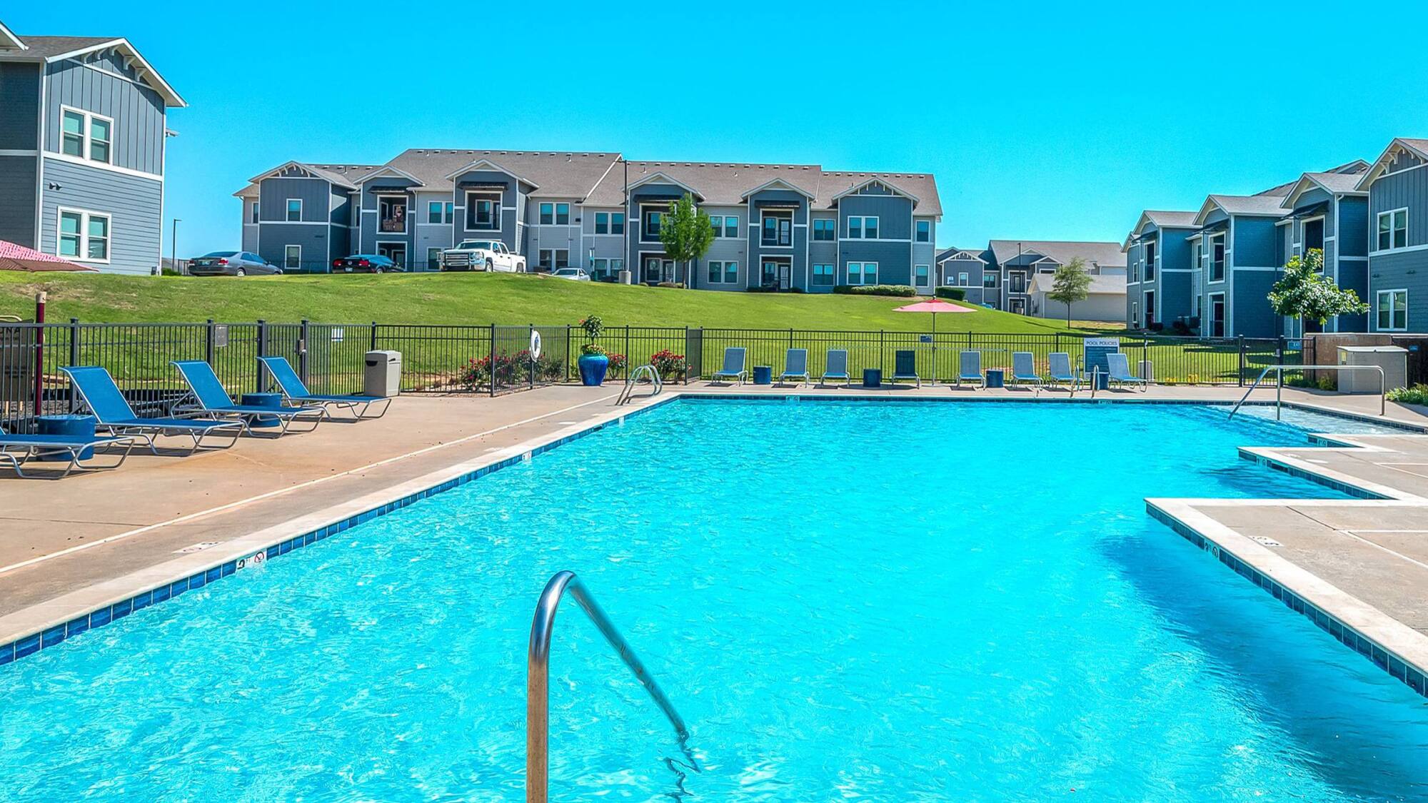 Outdoor swimming pool with lounge chairs, surrounded by modern apartment buildings on a sunny day.