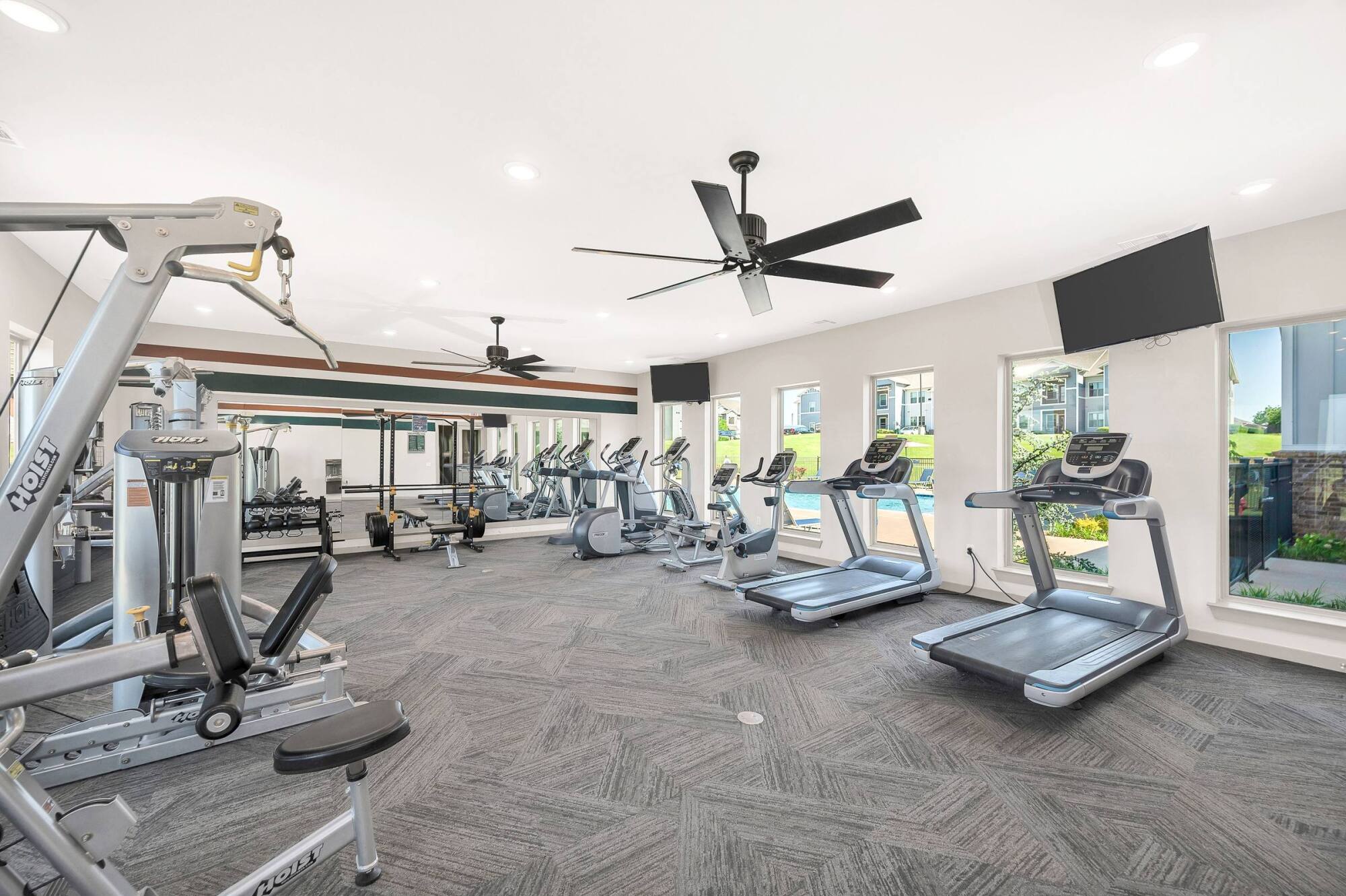 Modern gym with treadmills, exercise machines, free weights, large mirrors, TVs, and windows letting in natural light.