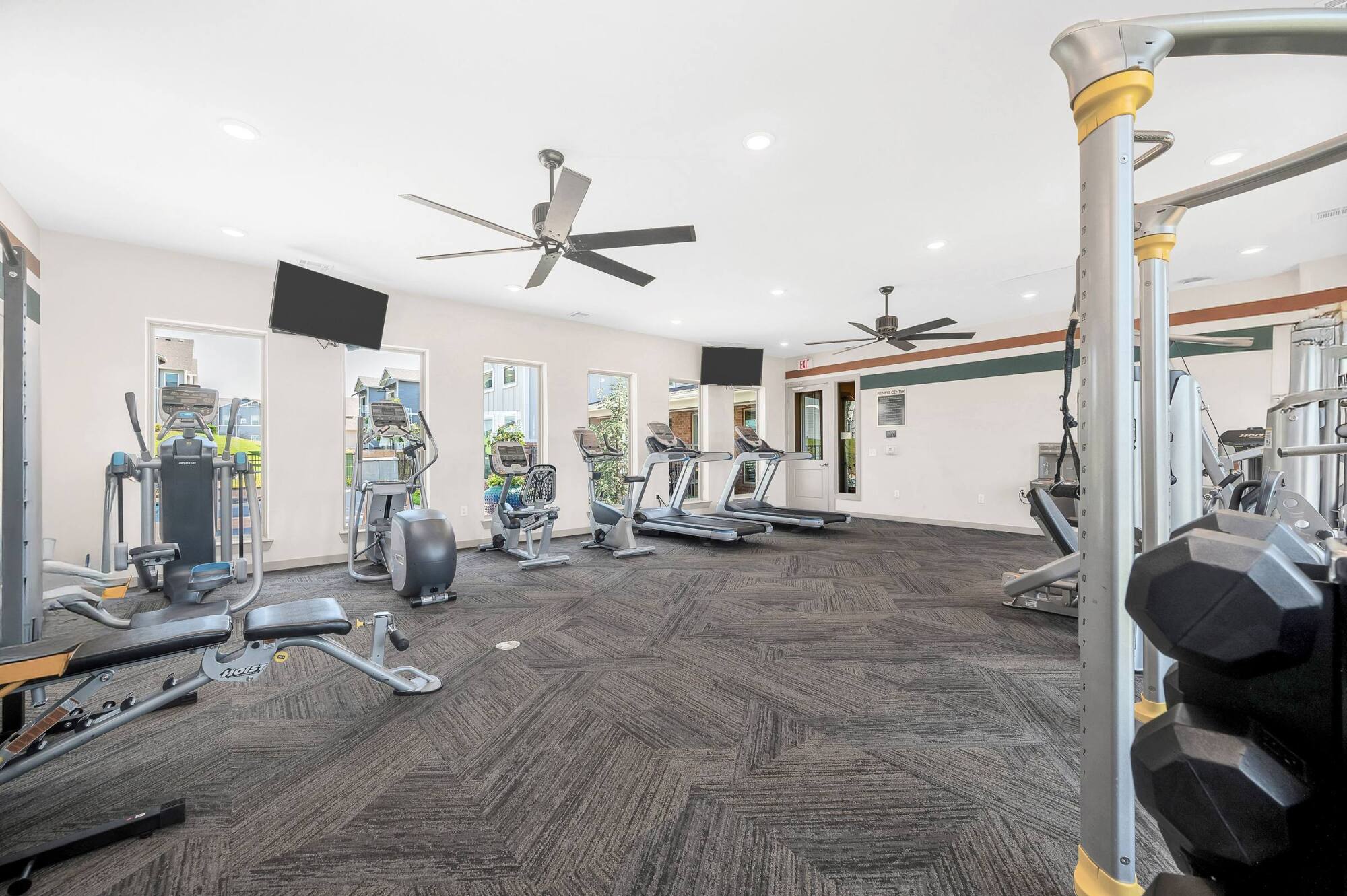 A bright gym with treadmills, exercise bikes, weight machines, free weights, and ceiling fans.