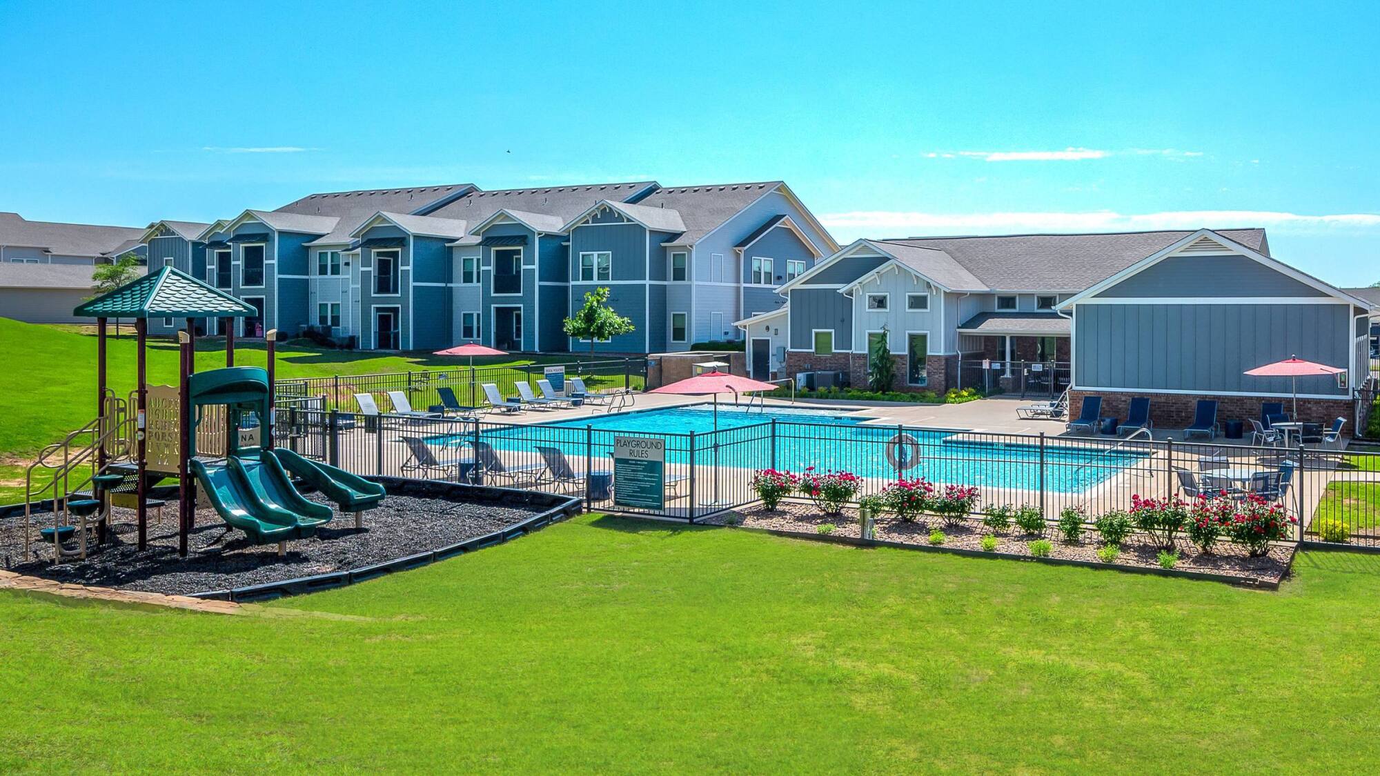 Modern apartment complex with a playground, fenced outdoor pool, lounge chairs, and landscaped green lawn.