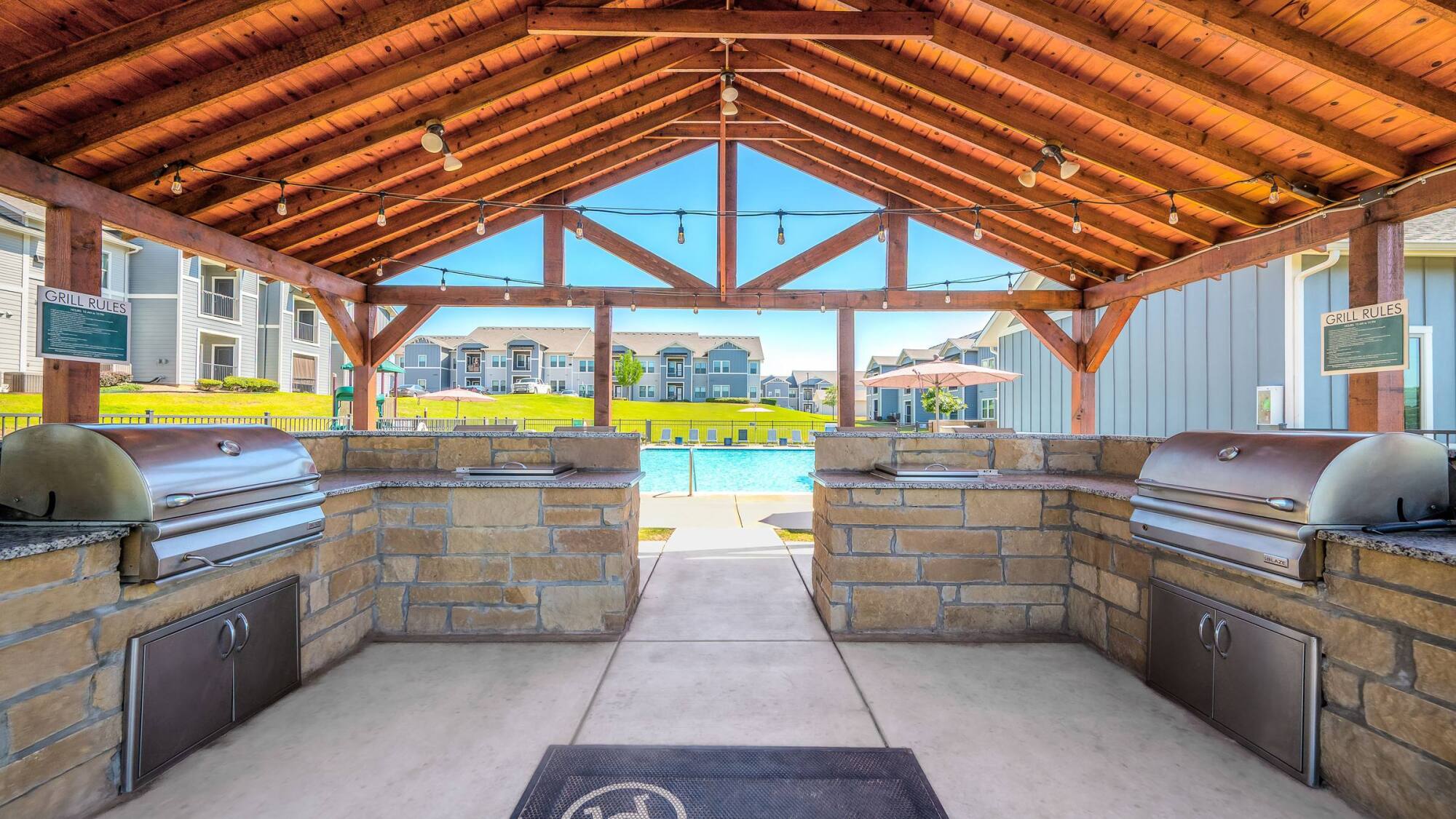 Outdoor covered grilling area with two grills, stone counters, and a view of an apartment pool and buildings.