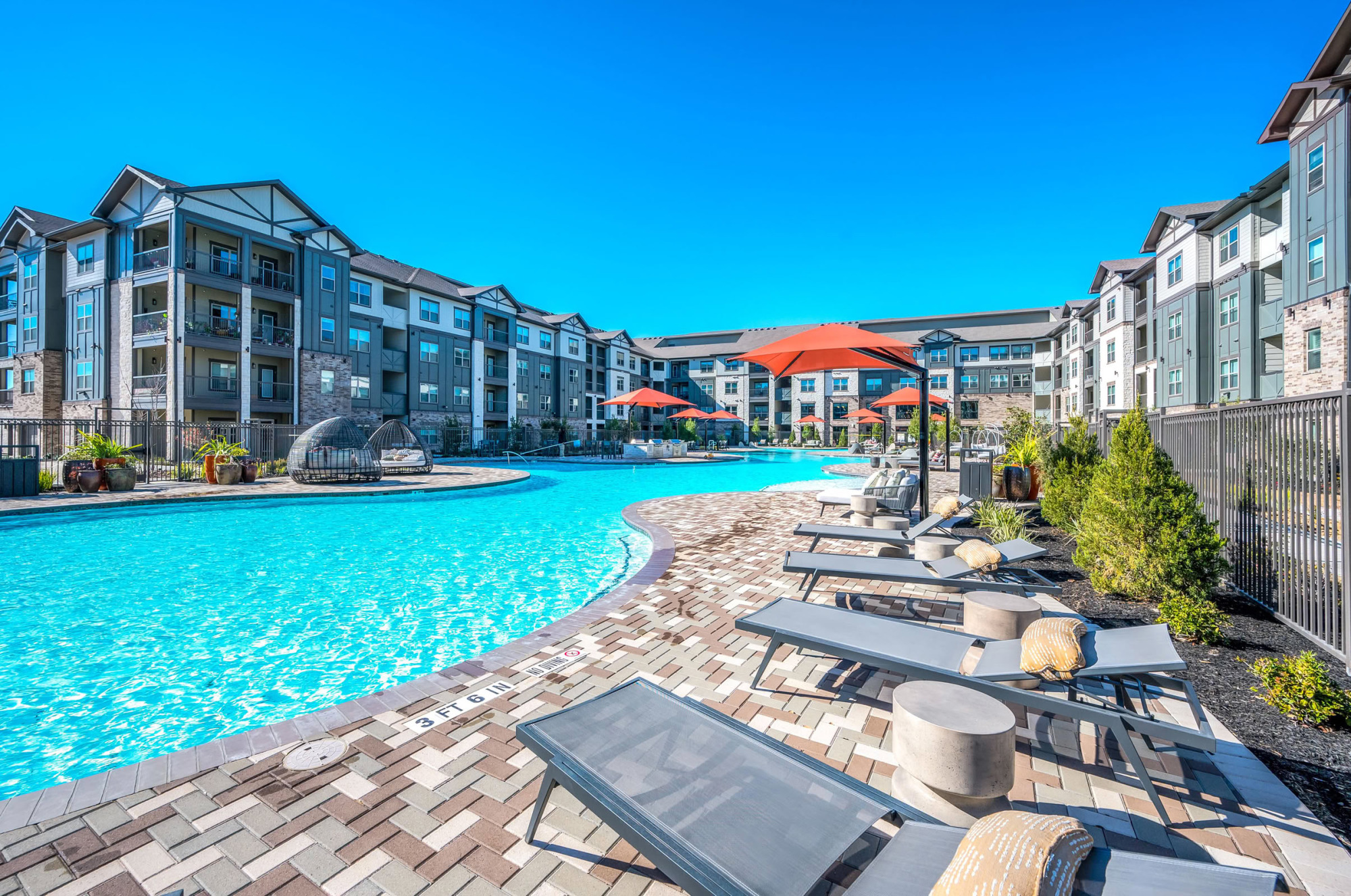 Curved resort-style pool lined with paver decking, lounge chairs, orange umbrellas, lush landscaping, and a multi-story apartment community visible against a vivid blue sky.