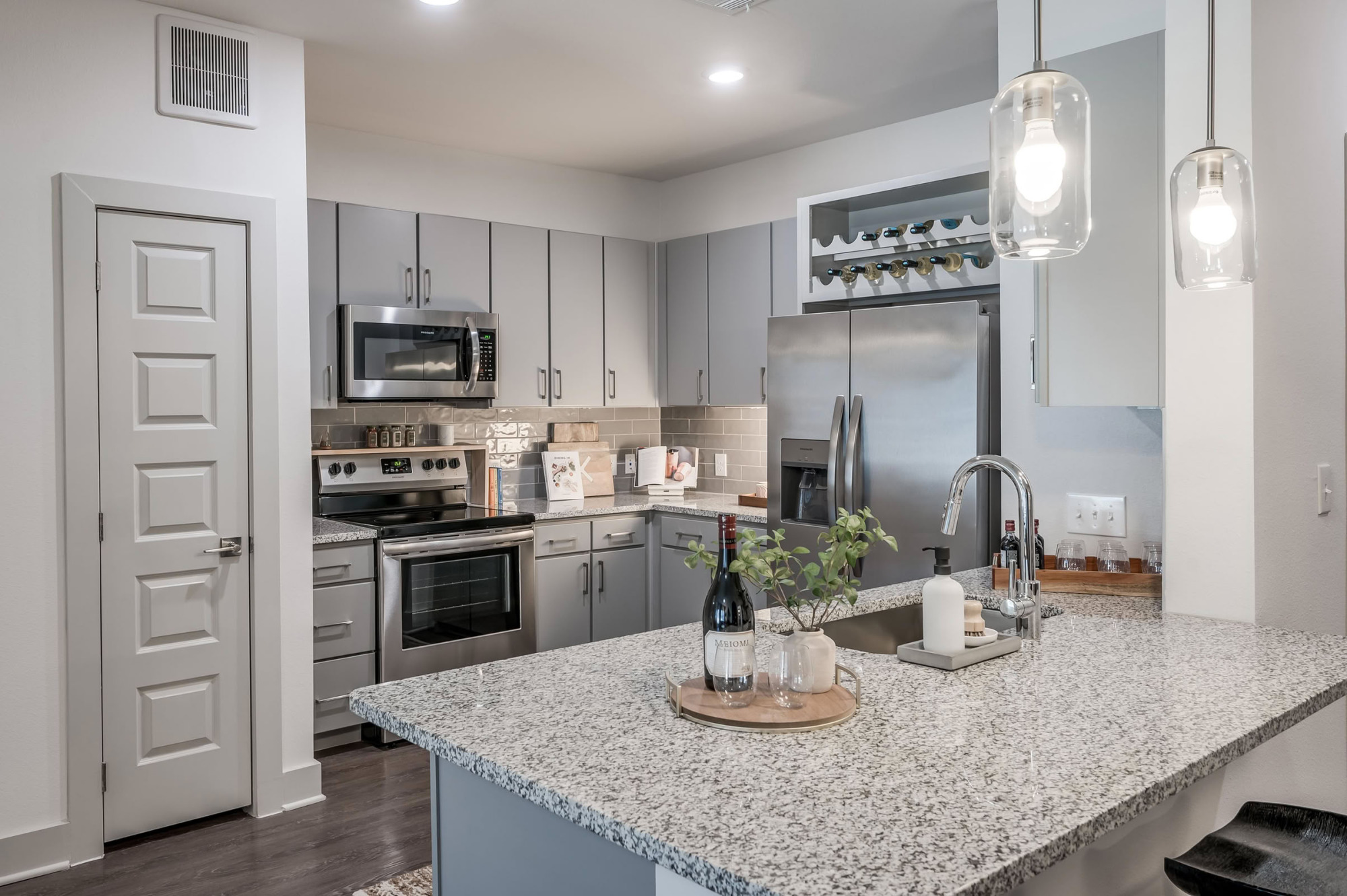 Stylish apartment kitchen with gray cabinetry, granite-topped peninsula, stainless steel appliances, built-in wine storage, and modern glass pendant lighting over the breakfast bar.