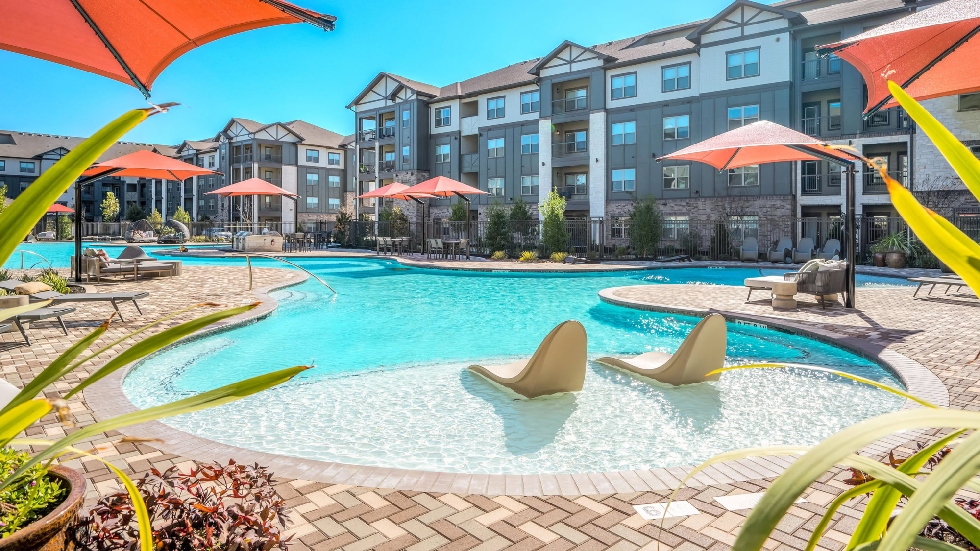 Sun-drenched community pool with in-water tanning chairs, vibrant orange umbrellas, paver patios, tropical plantings in the foreground, and modern apartment buildings surrounding the courtyard.