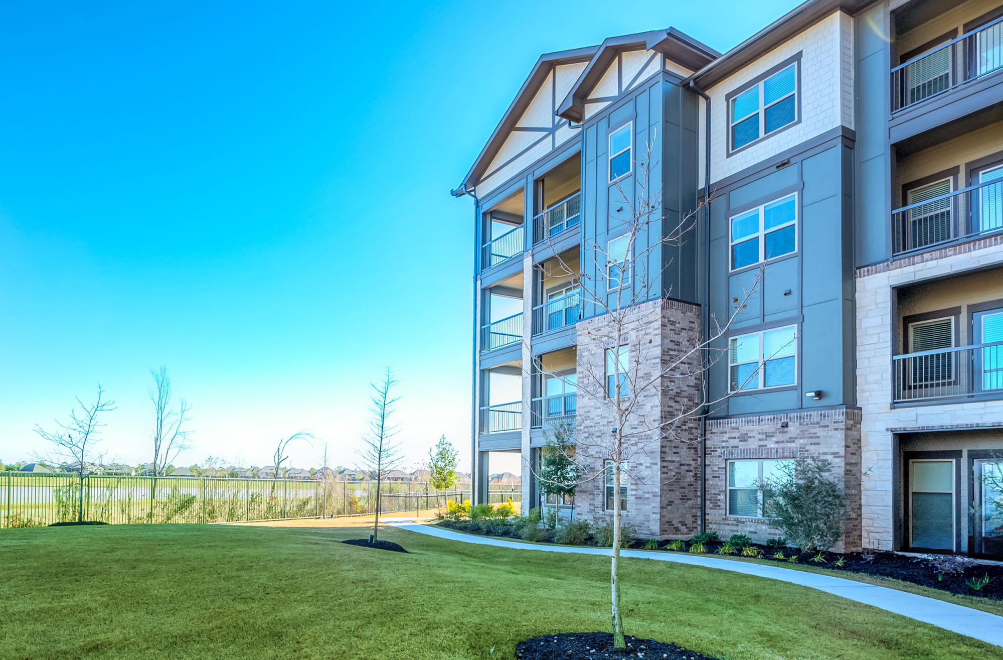 Exterior view of a four-story apartment building with mixed brick, stone, and gray siding facade, private patios and balconies, manicured lawn, and a young tree under a clear blue sky.