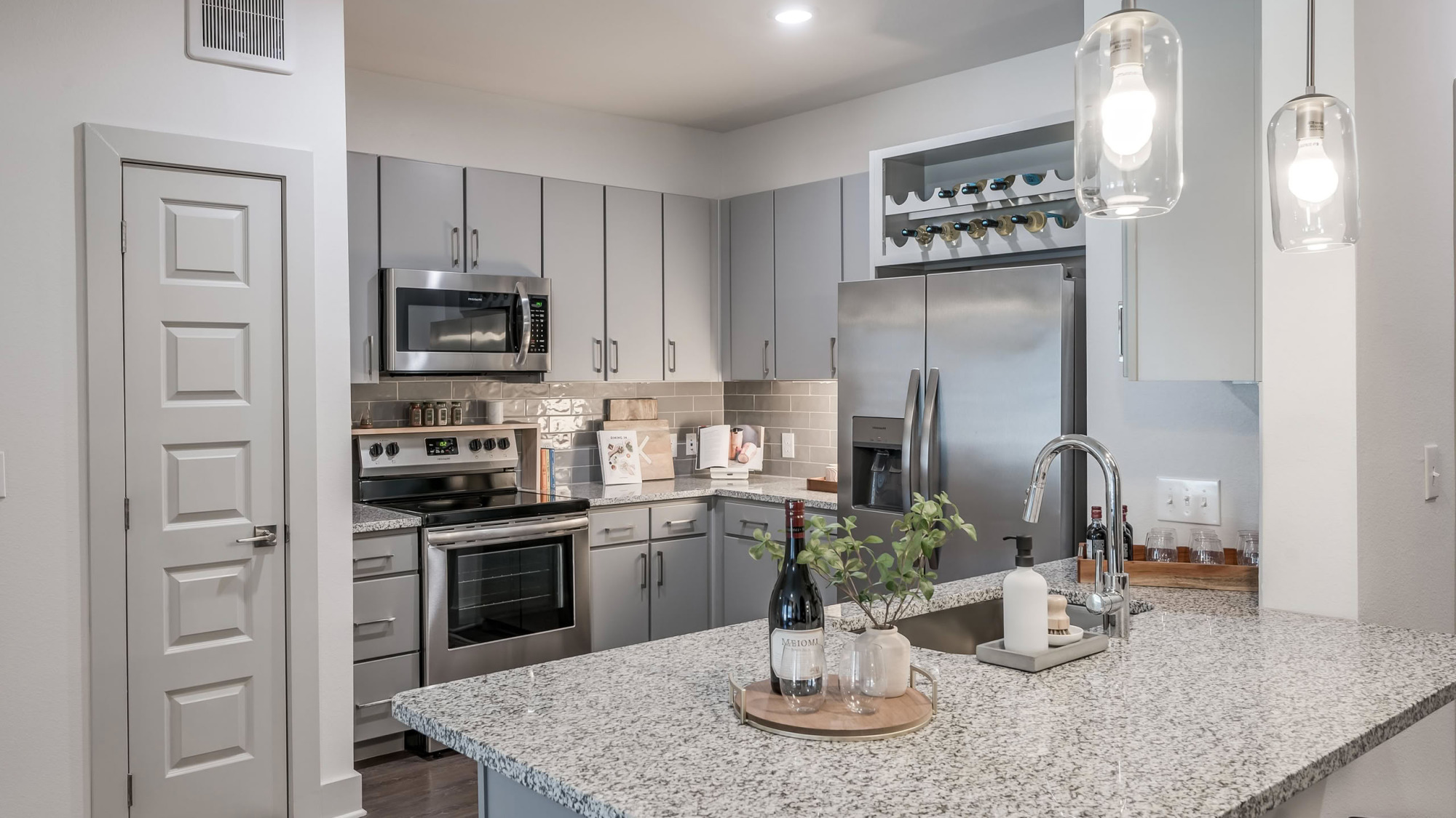 Contemporary apartment kitchen featuring light gray cabinetry, granite countertops, stainless steel appliances, a built-in wine rack, glass pendant lighting, and a subway tile backsplash.