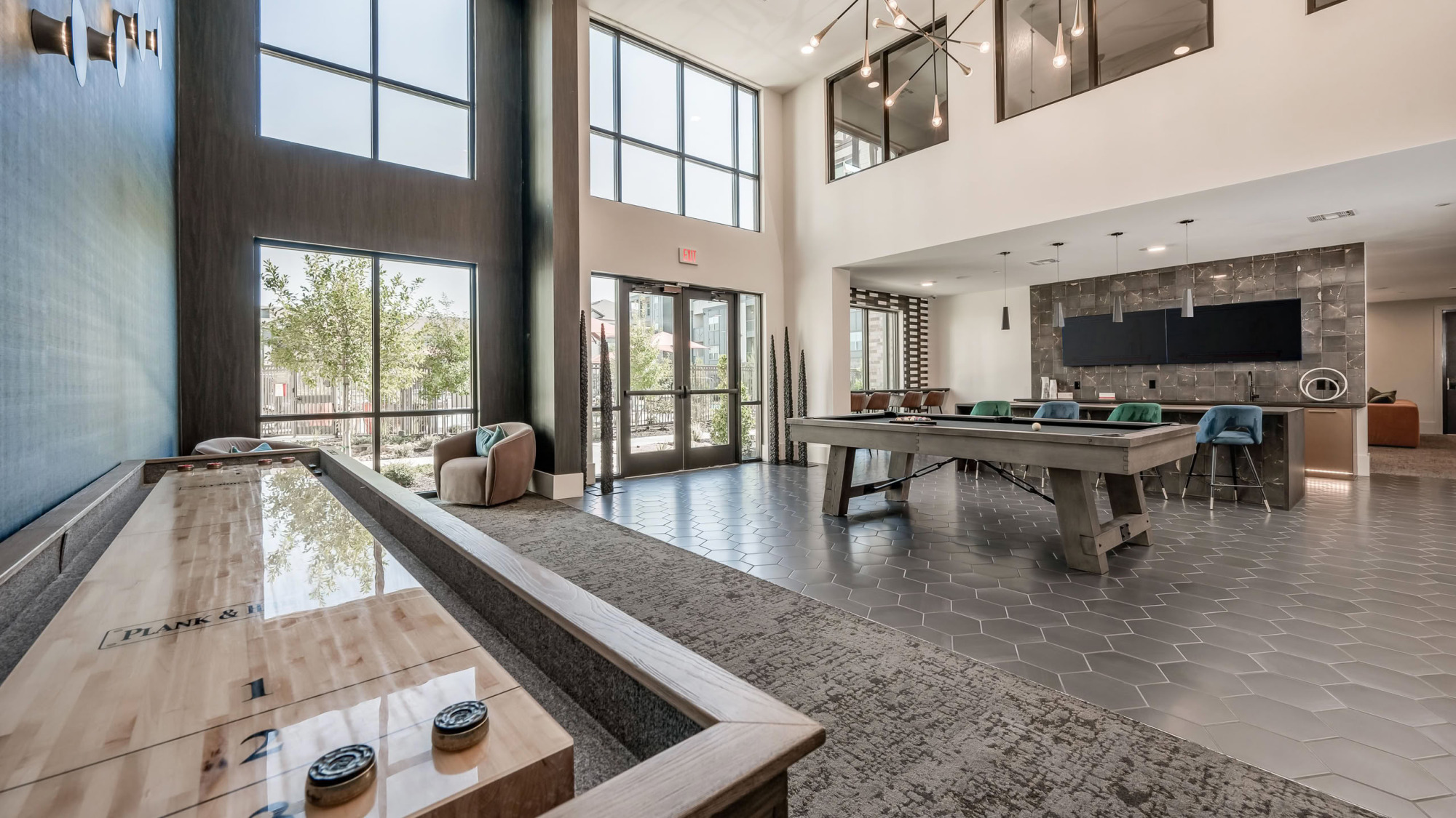 Upscale resident clubhouse lounge featuring a long shuffleboard table, billiards table, hexagonal tile flooring, two-story floor-to-ceiling windows, and modern statement chandeliers.
