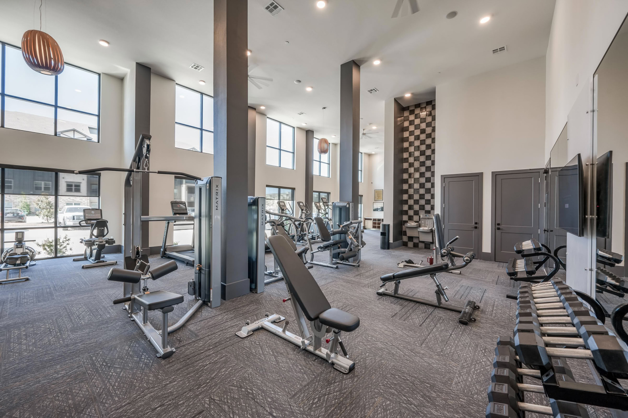 Spacious two-story gym featuring strength training machines, an adjustable bench, treadmills by floor-to-ceiling windows, and a stylish checkered tile accent wall.