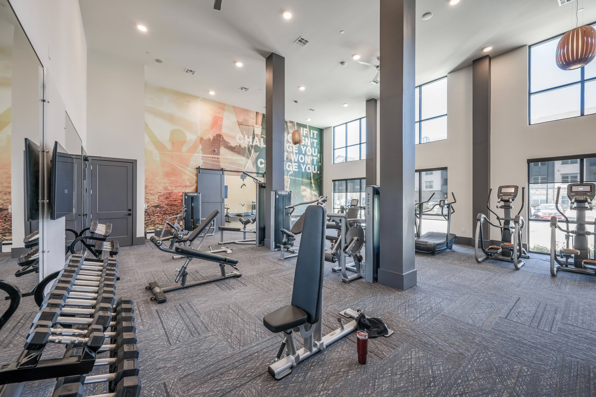 Modern fitness center with high ceilings, weight machines, free weights, a bench press station, cardio equipment, and motivational graphic wall art lining the wall.