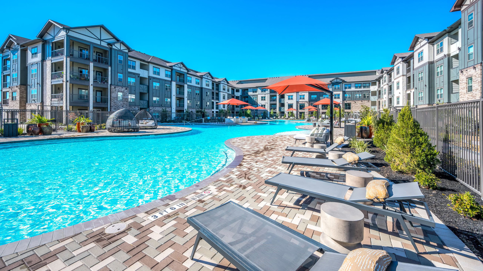Resort-style swimming pool surrounded by gray and orange umbrellas, lounge chairs, paver stone decking, and a modern multi-story apartment community in the background under a clear blue sky.