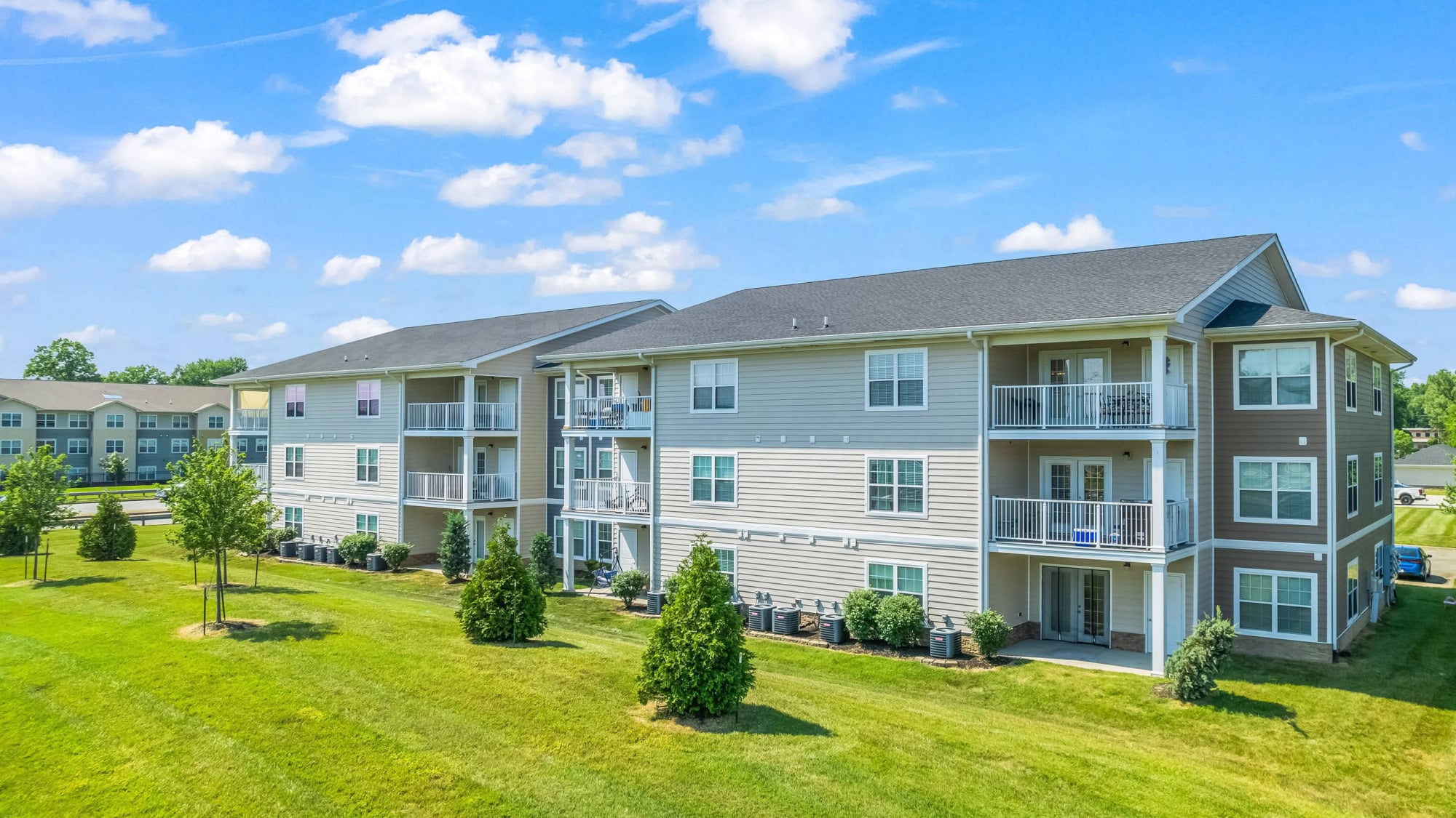 Three-story tan-sided apartment building with white-railed balconies, a peaked roofline, manicured landscaping, and a sweeping green lawn under a partly cloudy sky.
