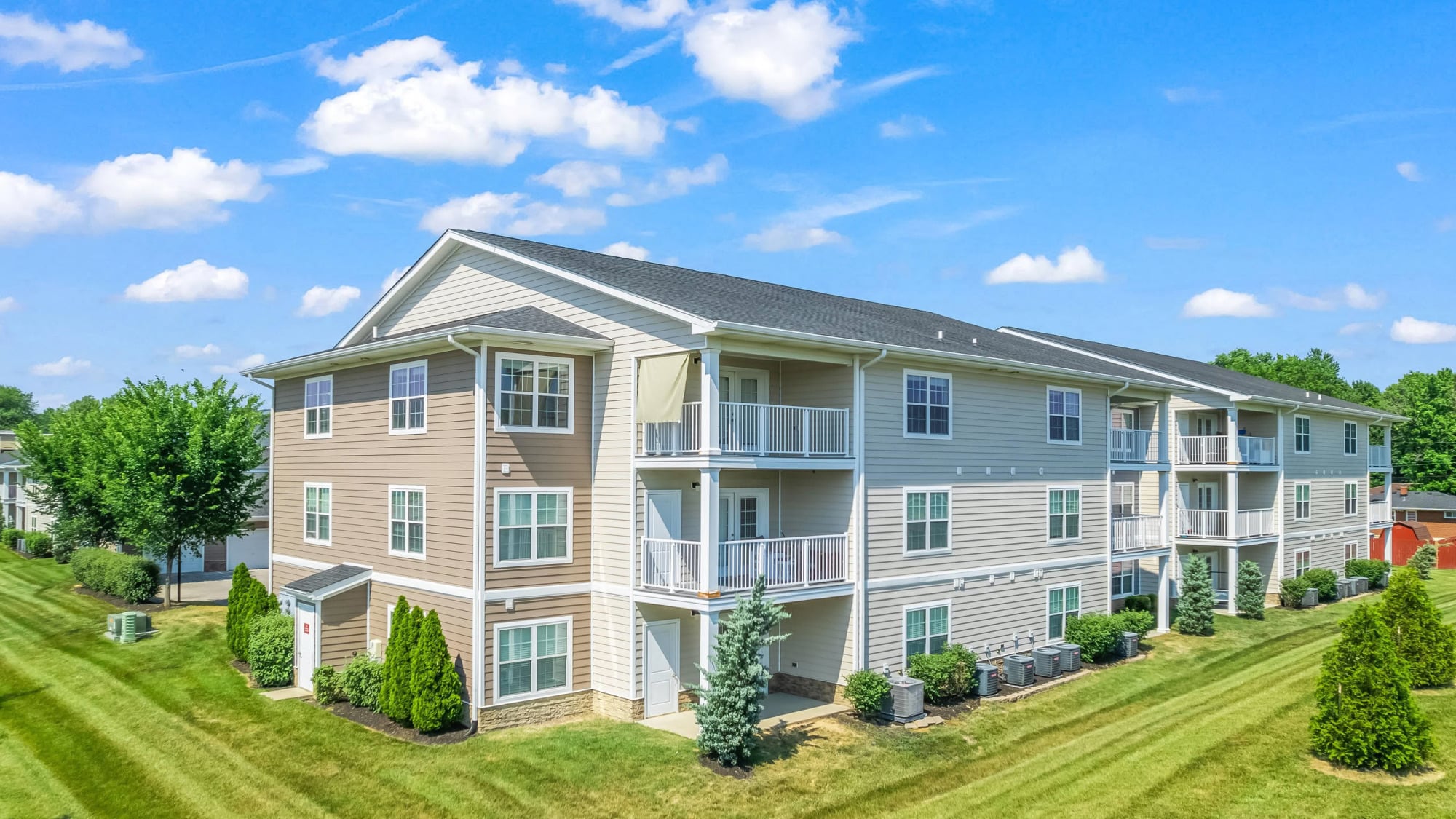 Three-story tan and white apartment building with private balconies, white trim, manicured shrubs, and a rolling green lawn under a blue sky with scattered clouds.