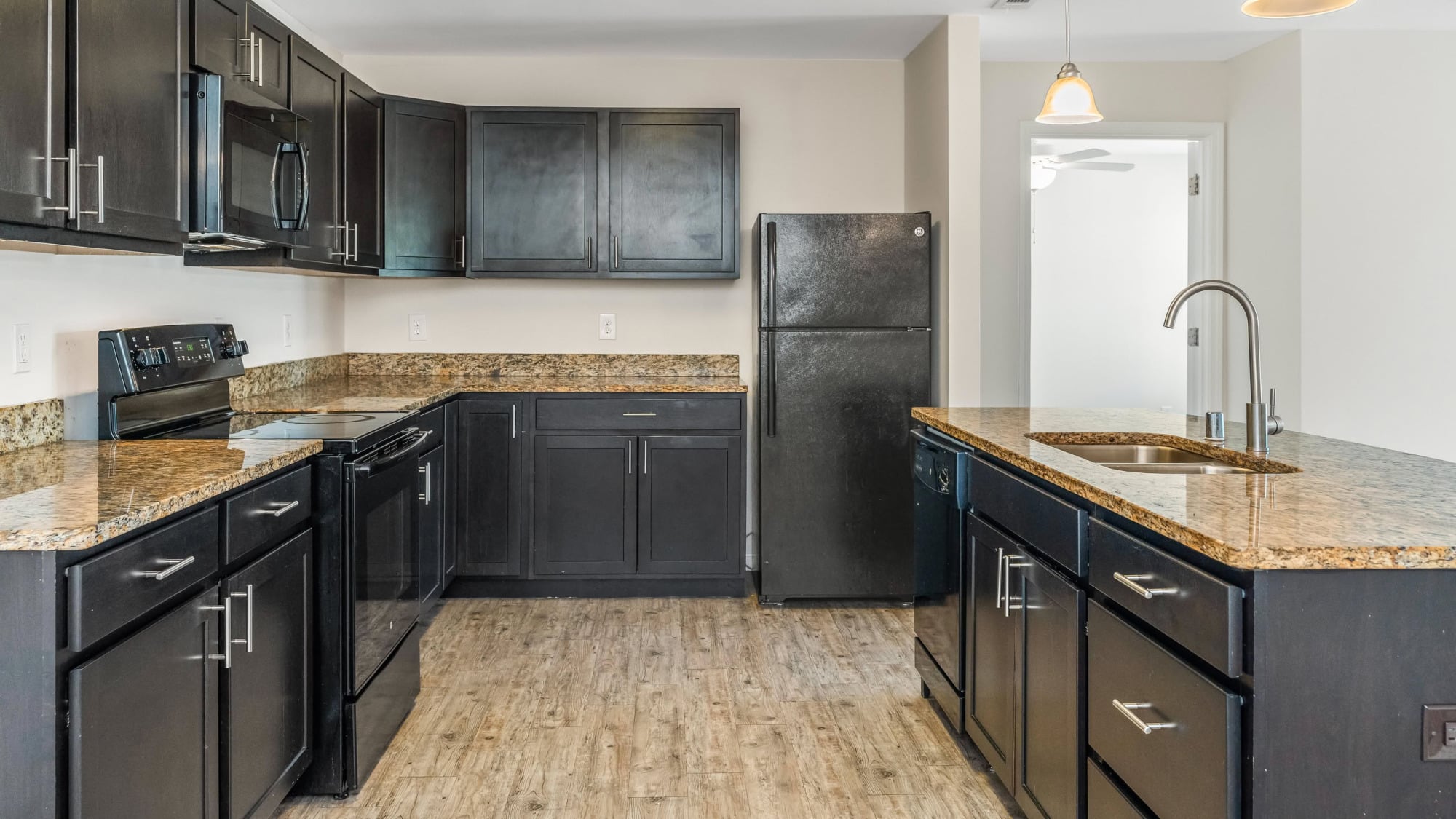 Apartment kitchen with espresso-stained shaker cabinets, granite countertops, black appliances including a stove and refrigerator, light wood-look flooring, and a pendant light over the breakfast bar.