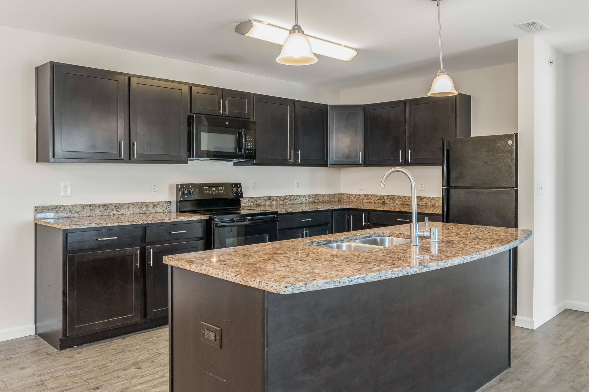 Spacious apartment kitchen featuring dark espresso cabinetry, granite countertops, a center island with double sink, black appliances, and dual pendant lights illuminating the workspace.