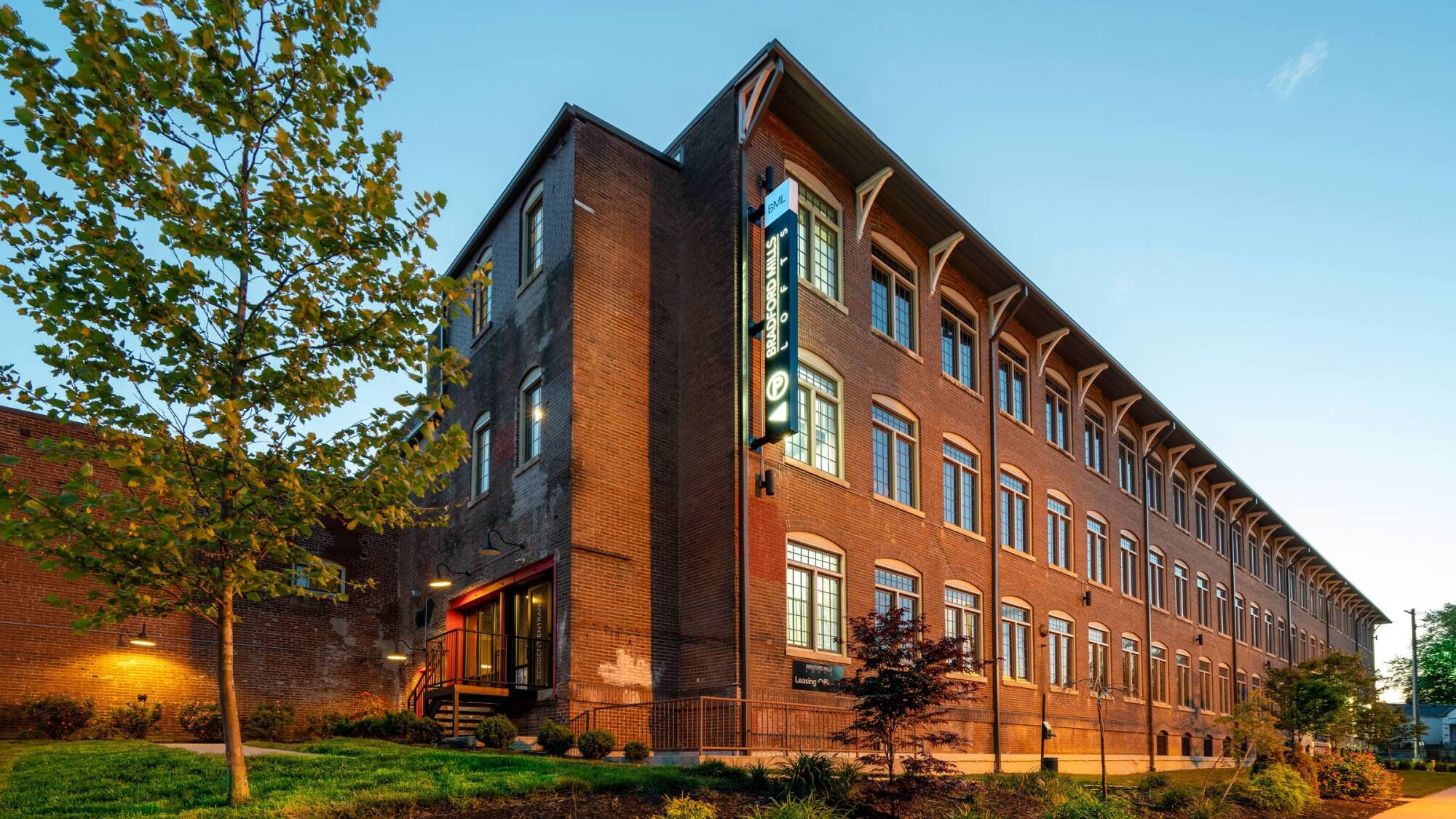 A large brick building with tall windows and a lit vertical sign, surrounded by trees at sunset.