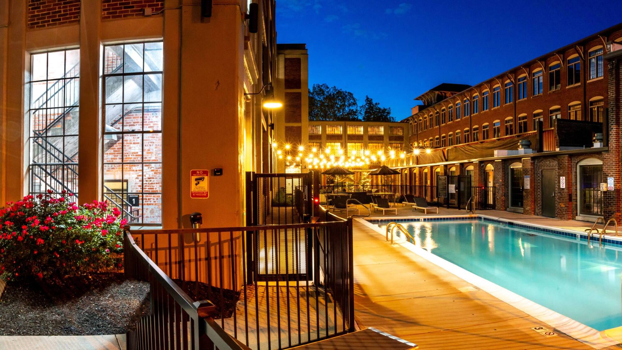 Nighttime view of a lit outdoor pool with lounge chairs and string lights next to a brick apartment building.