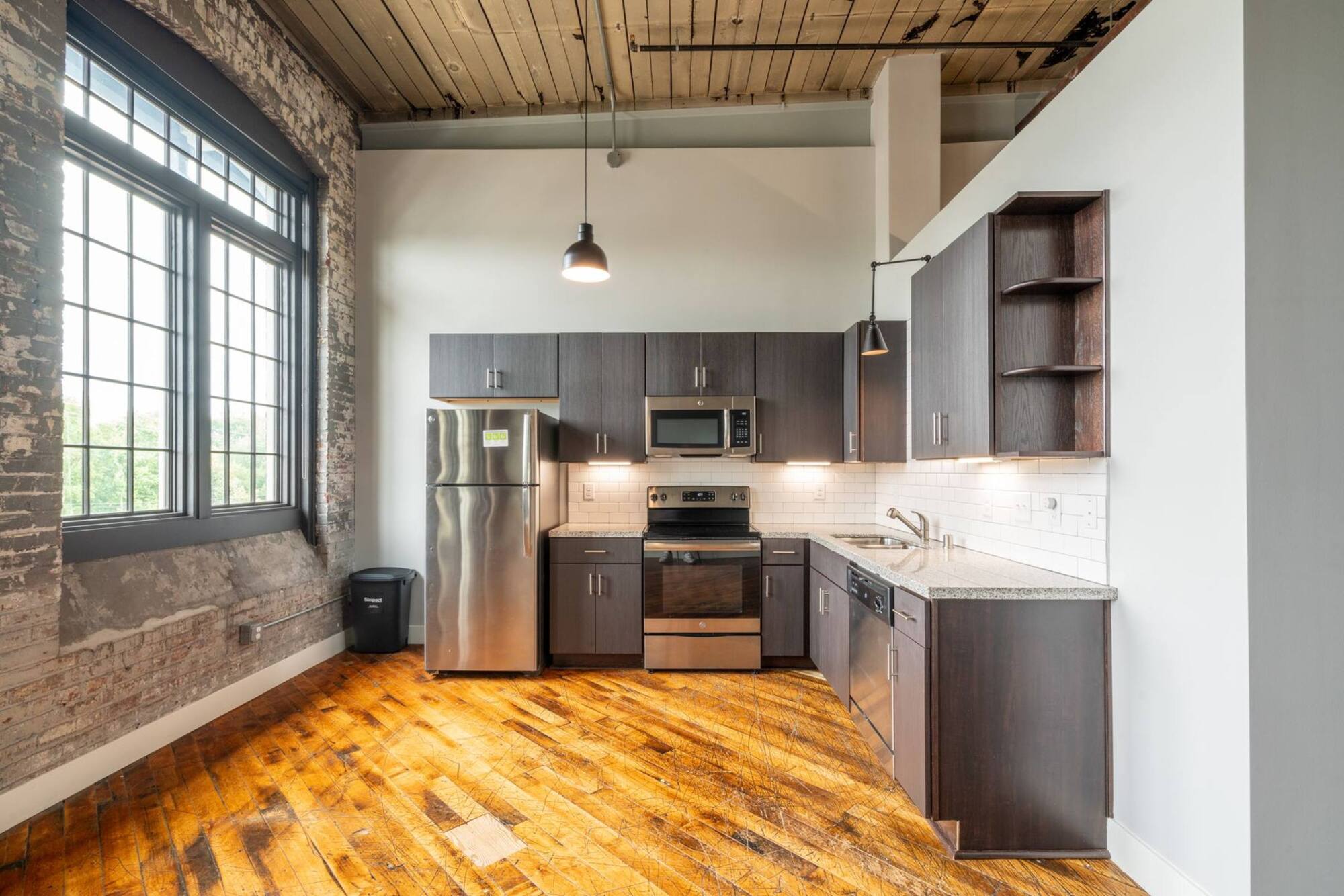 Modern kitchen with dark cabinets, stainless steel appliances, large window, and rustic wood floor.