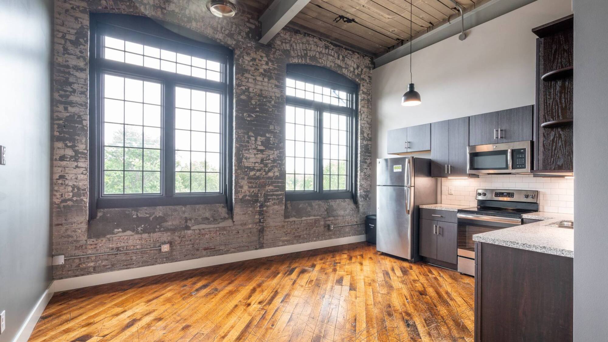 Loft kitchen with large industrial windows, exposed brick, wood ceiling, and hardwood floor.
