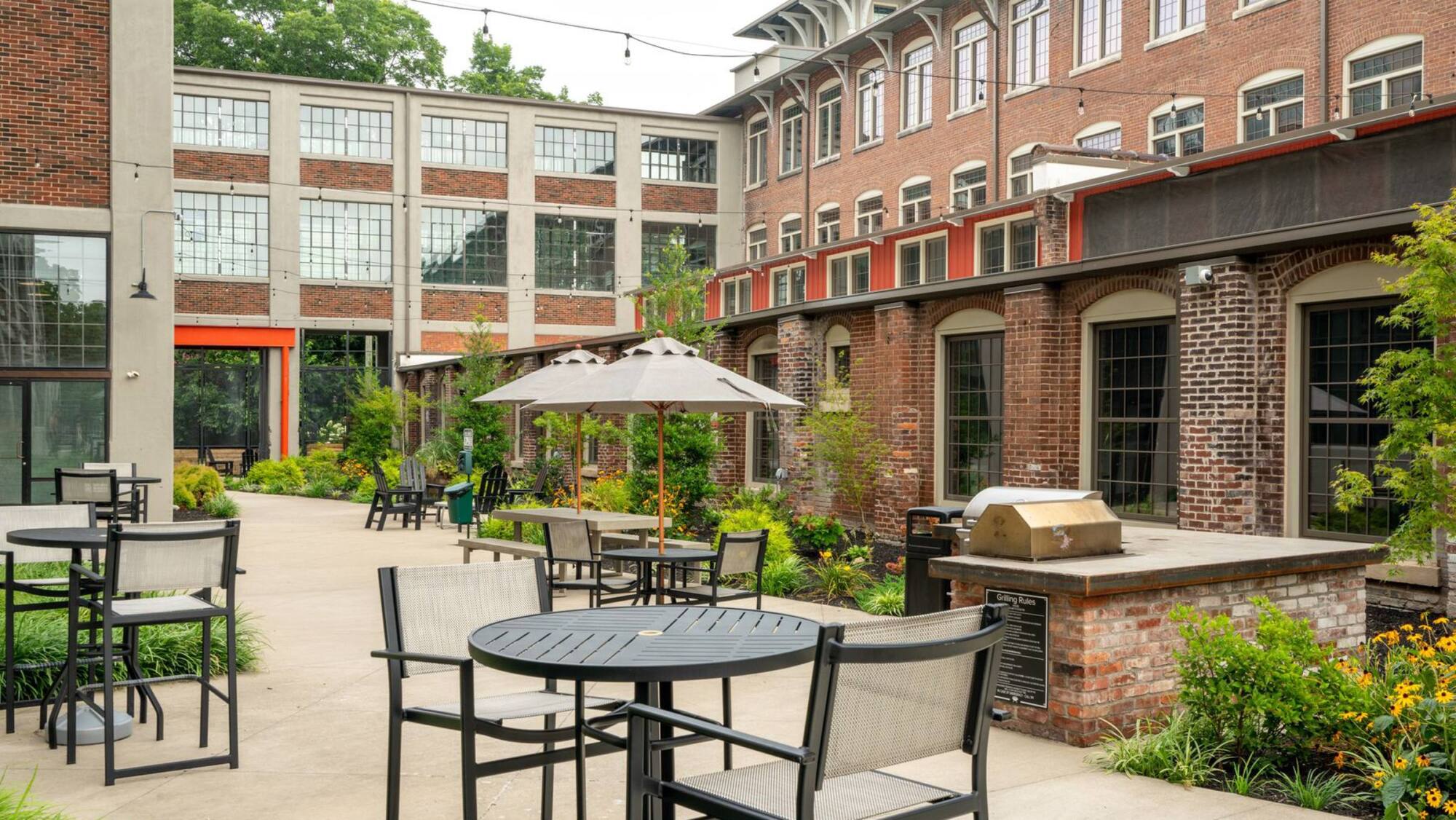 Outdoor patio with tables, chairs, umbrellas, a grill, and surrounding greenery beside brick buildings.