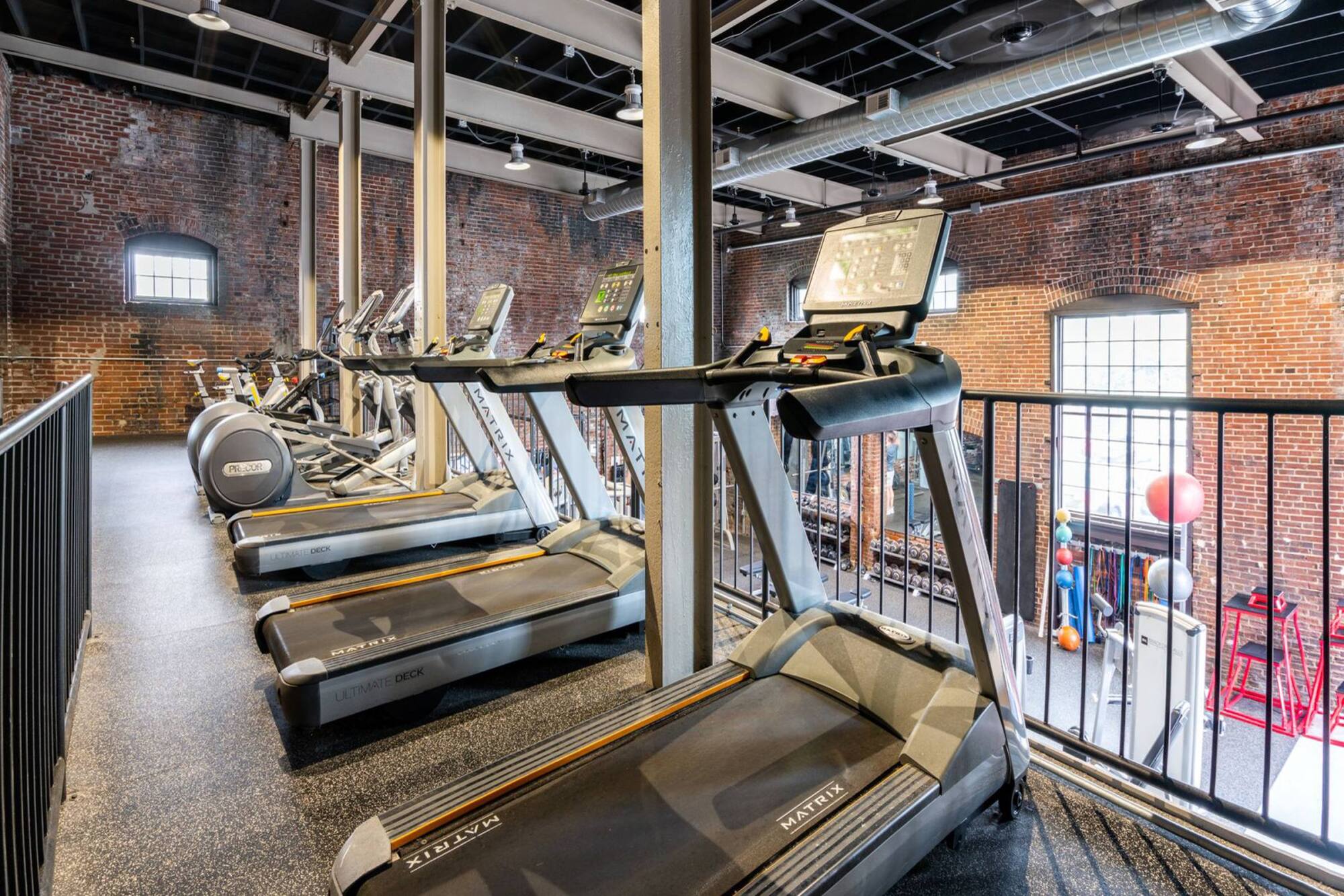 Treadmills and elliptical machines in a gym with brick walls and large windows, viewed from an upper level.
